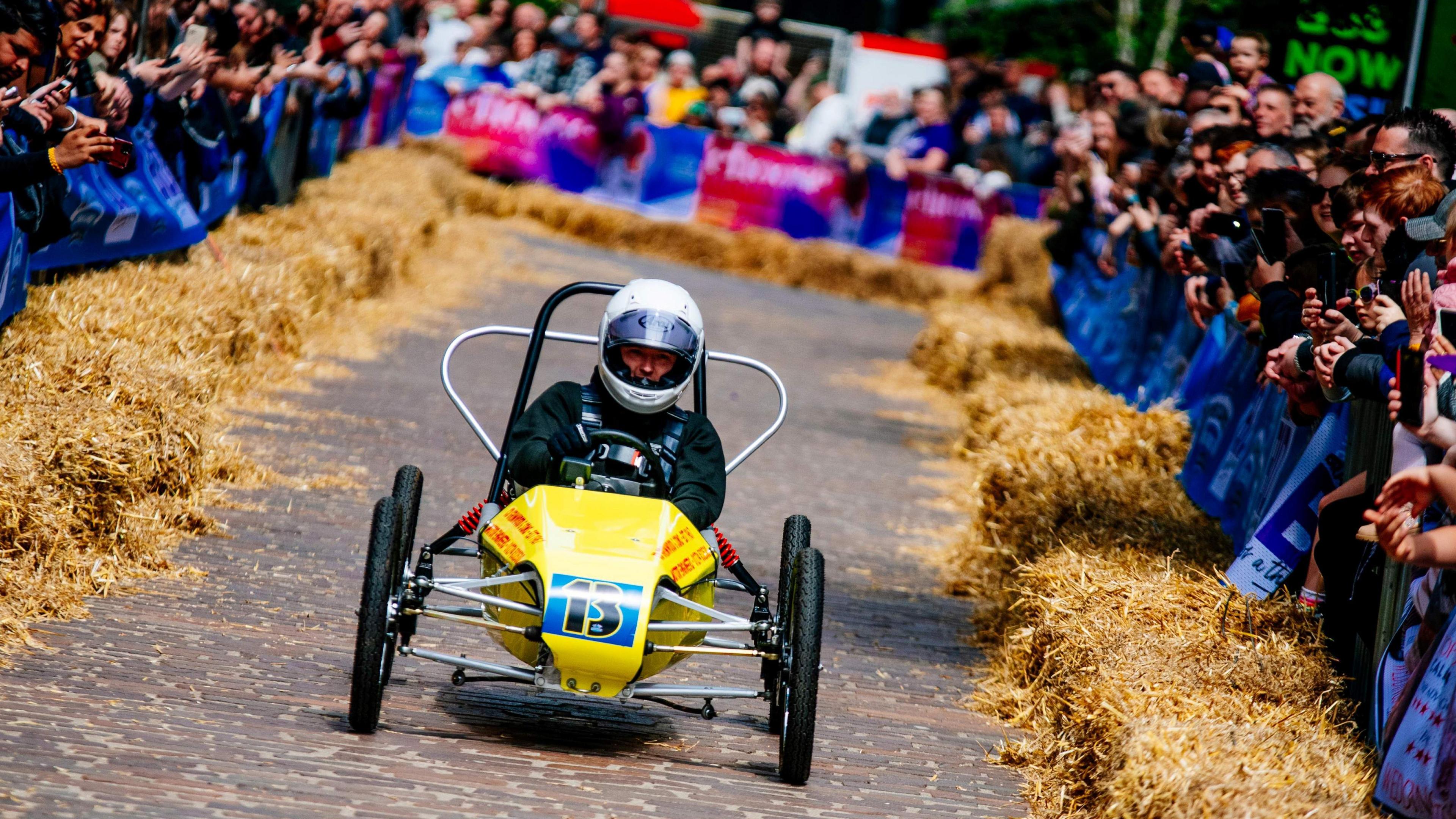 A go-kart with a driver wearing a crash helmet drives along a cobbled street with spectators either side behind hay bales.