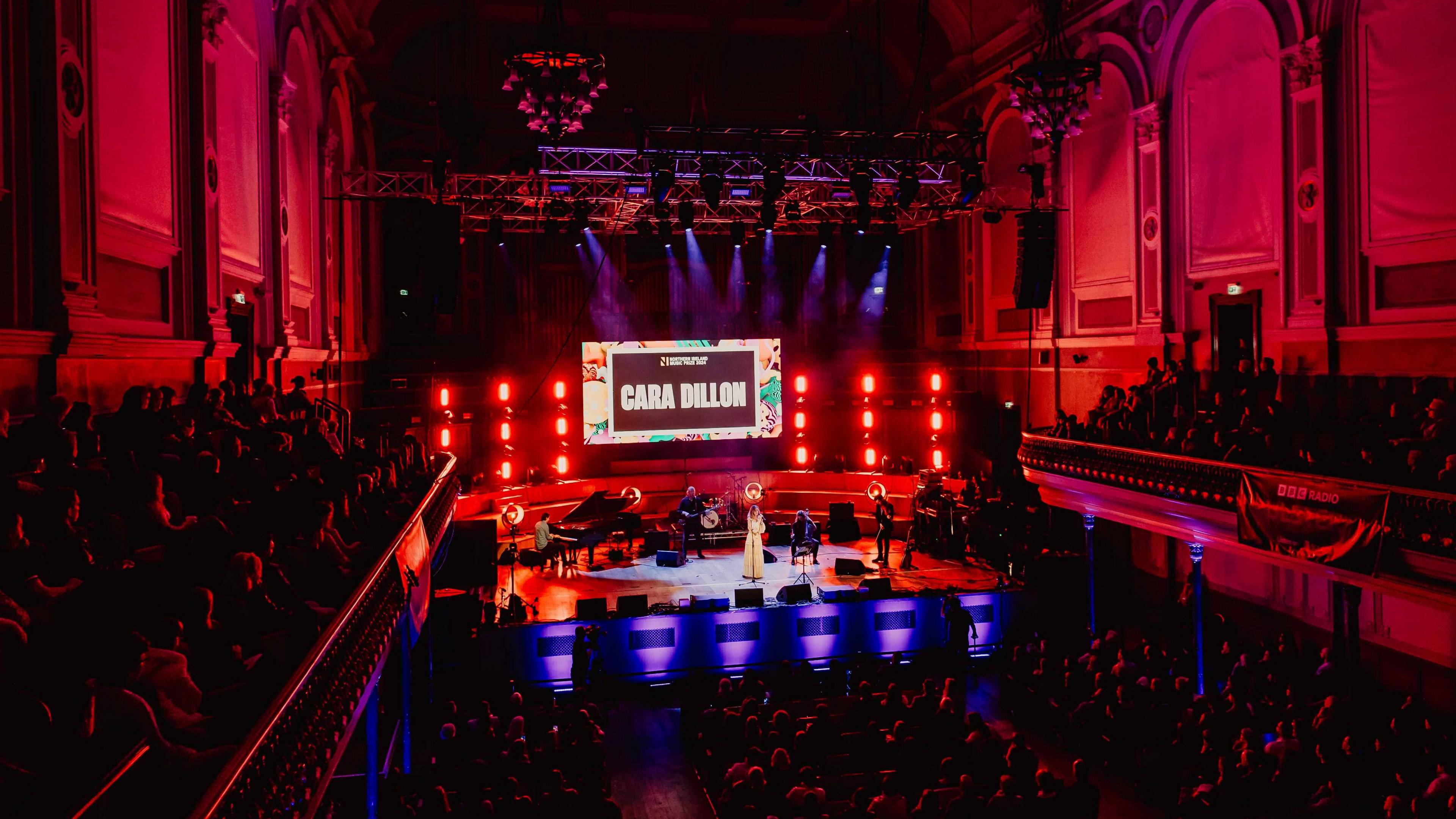 A large hall with music being played on stage with red lighting. 