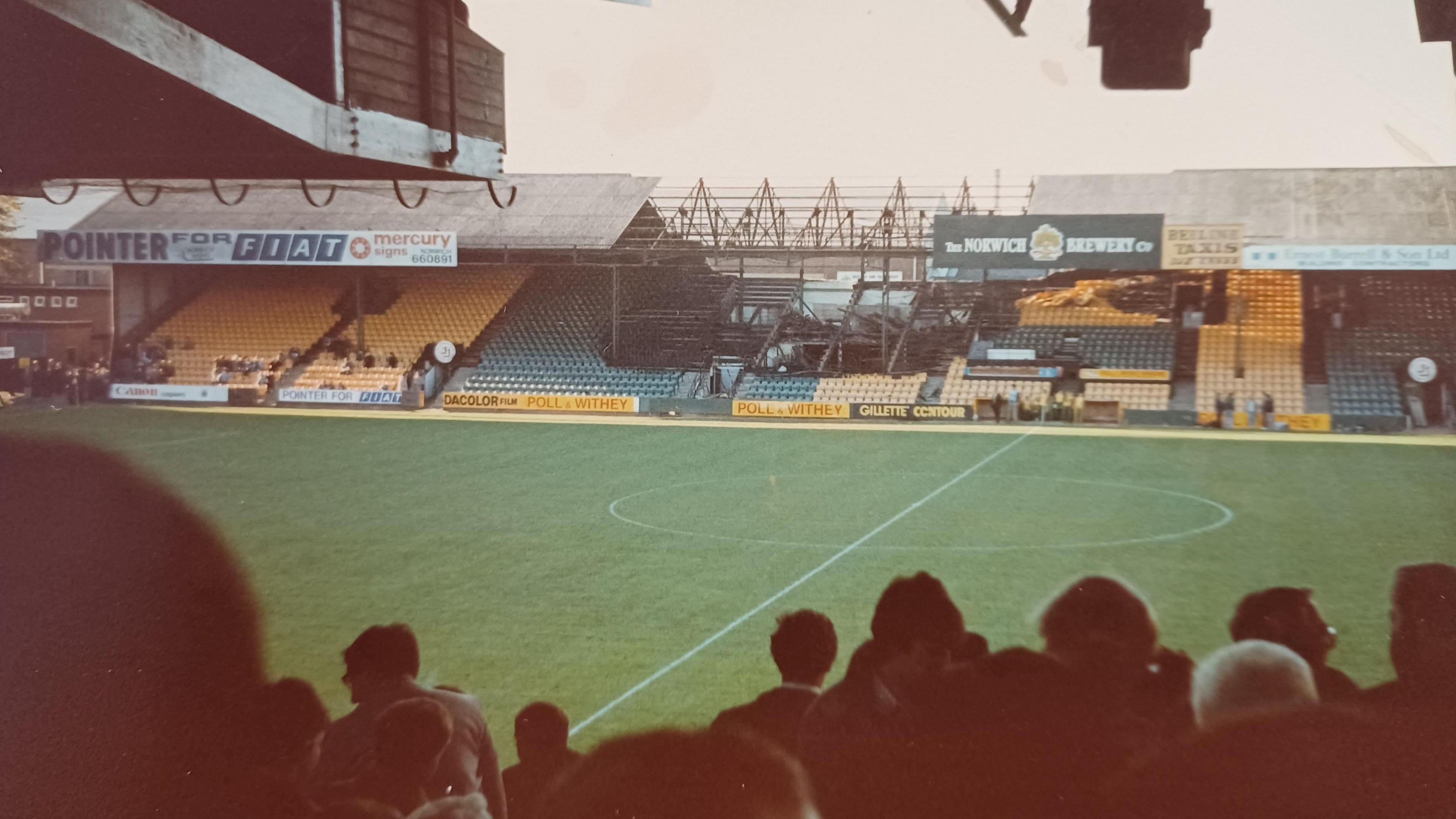 A fire-damaged Norwich City Football Club. You can see fans looking at a football pitch, a burned-out section of it and football signage.