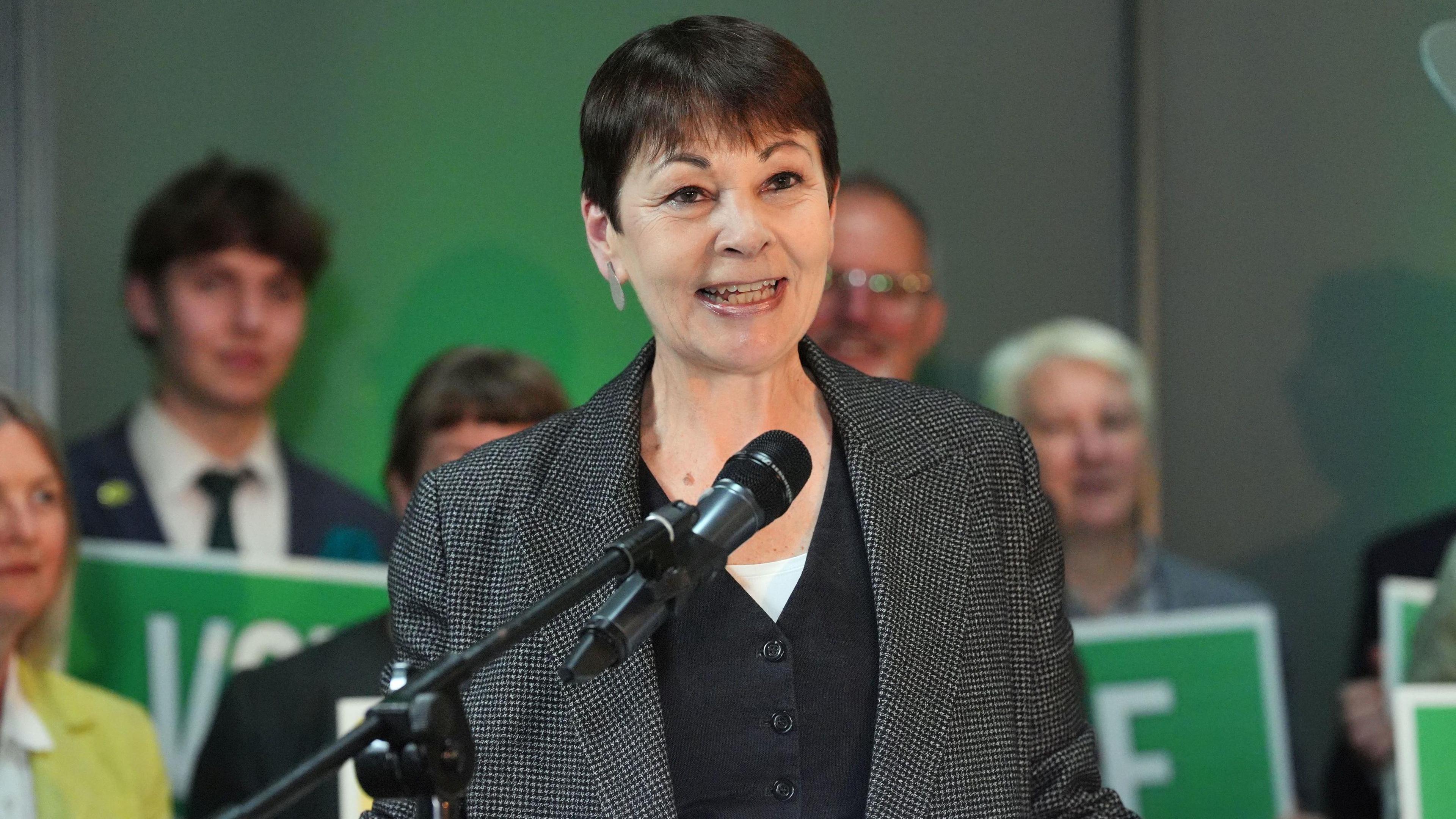 Caroline Lucas stands behind a microphone. She has a tweed-like jacket on top of a dark grey cardigan. She has short brown hair and stands in front of people holding green placards.