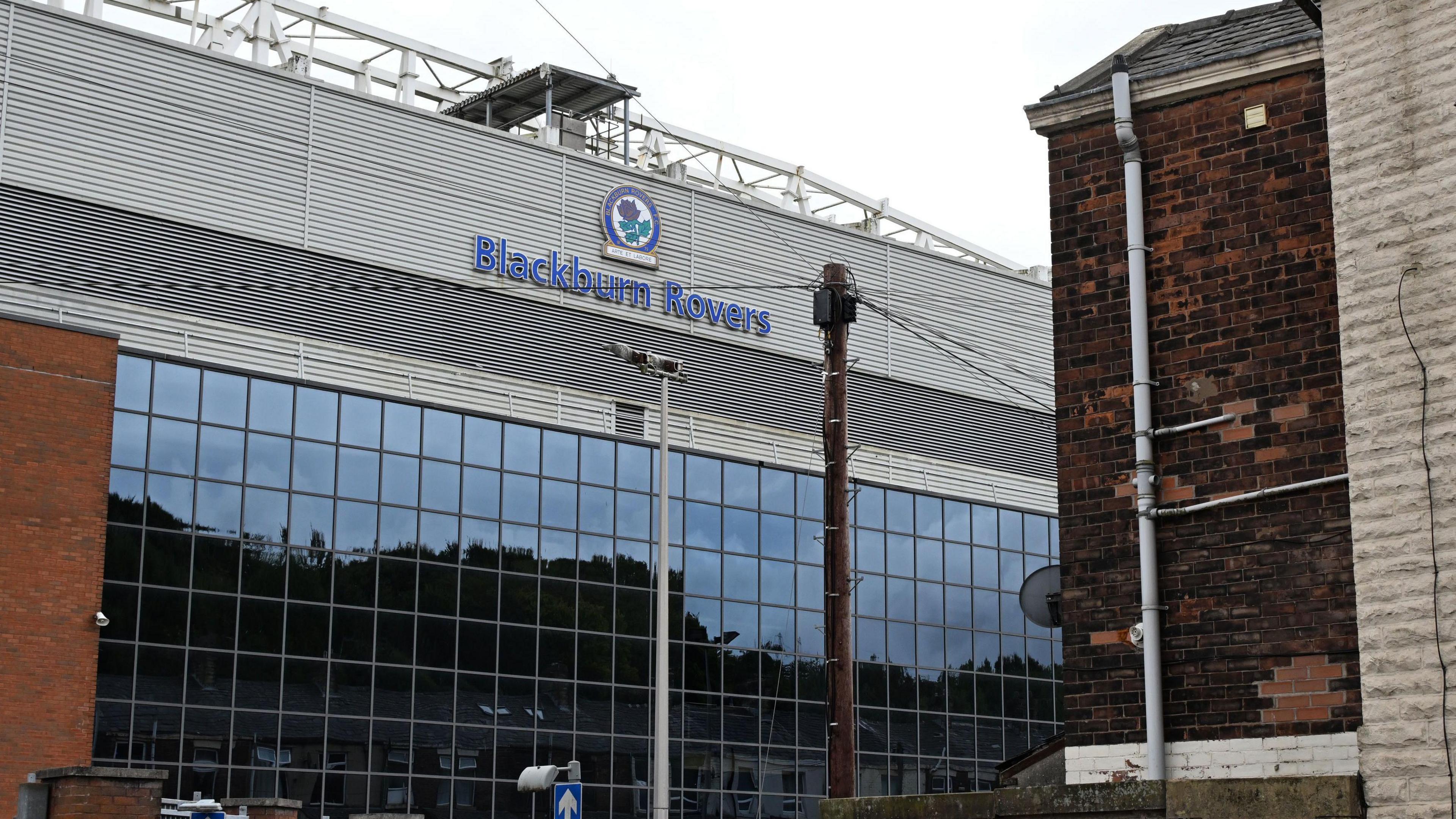 The outside of Ewood Park, showing a glass-fronted stand with the club logo of a red rose surrounded by a blue ring.