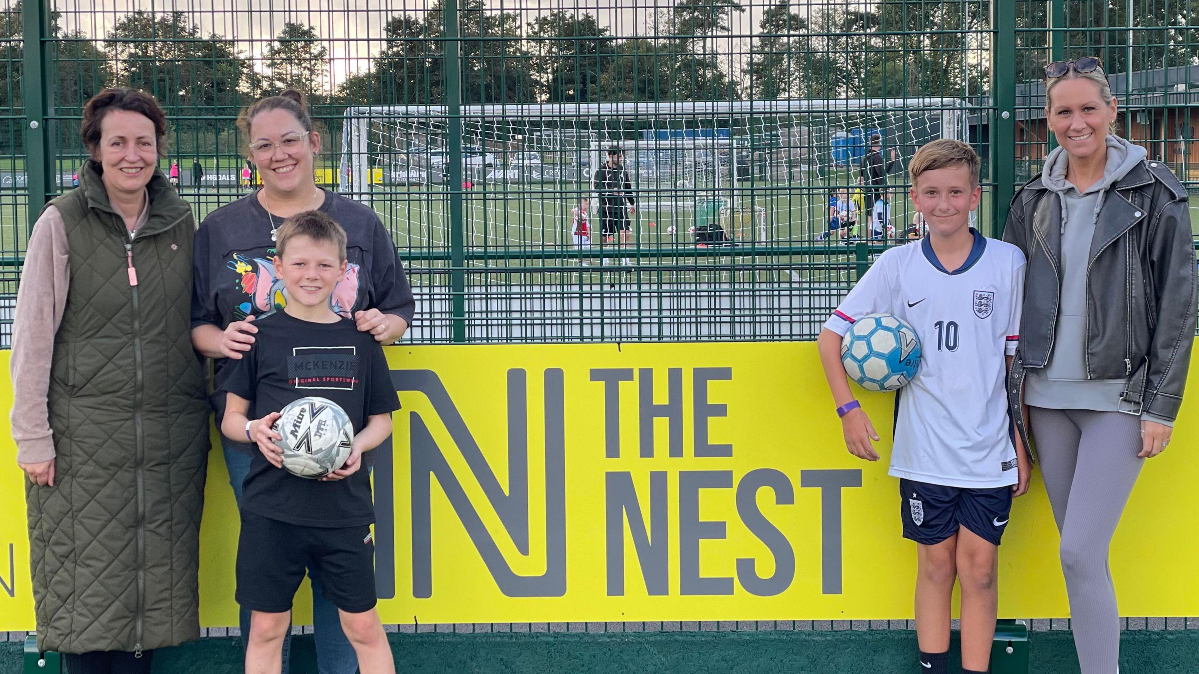 Three women and two boys holding footballs standing in front of a football pitch