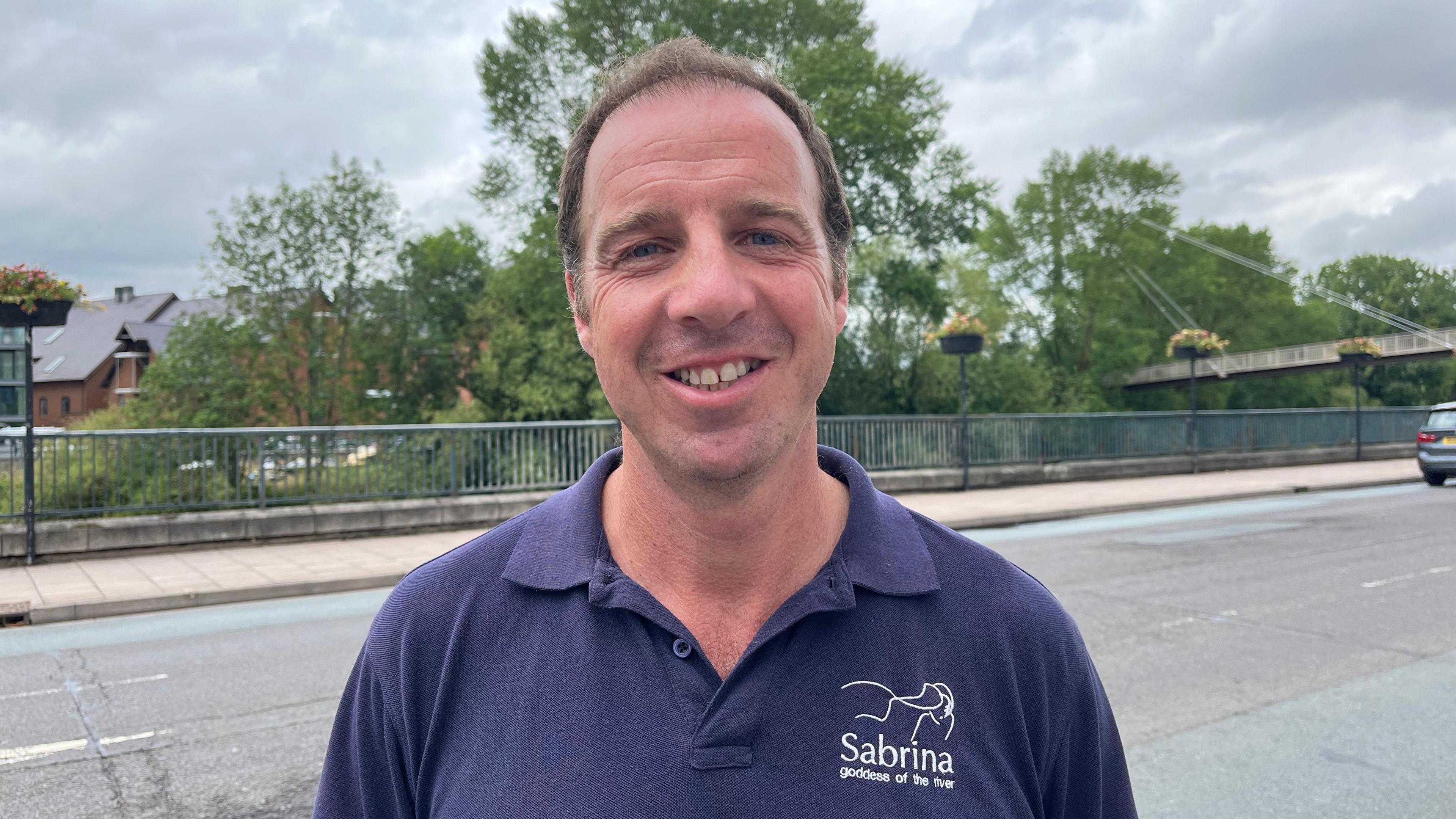 Dilwyn pictured smiling and looking into the camera. He's wearing a navy blue polo shirt with white writing that says 'Sabrina - goddess of the river.' He's stood by a road with a bridge overlooking the Severn.