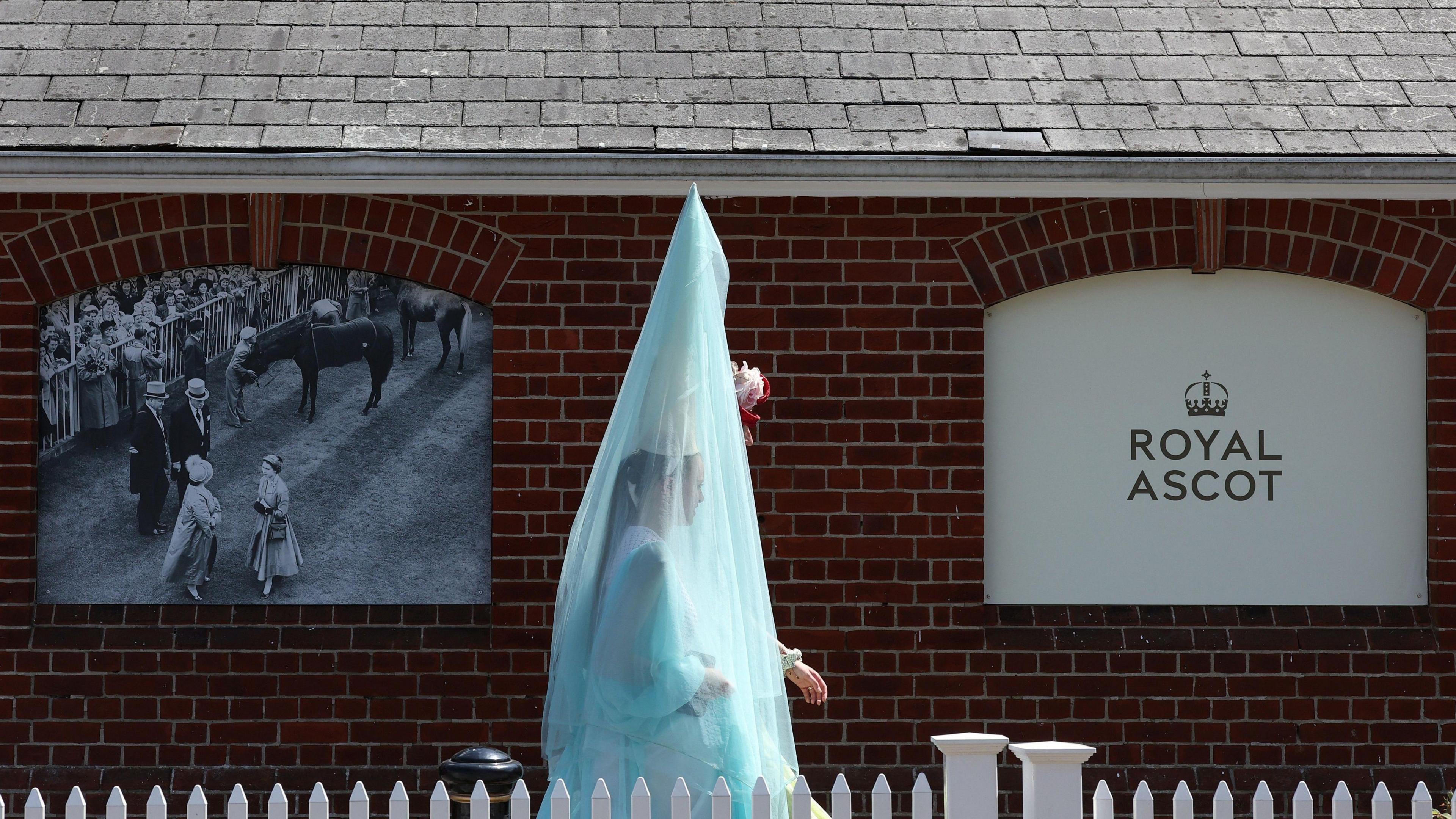 A woman walks past a red brick building, which has a black and white image of Queen Elizabeth II attending Royal Ascot on the left and a white sign which reads Royal Ascot on the right. She wears a tall pointed hat, which has a large turquoise veuil draped over it, covering her completely.