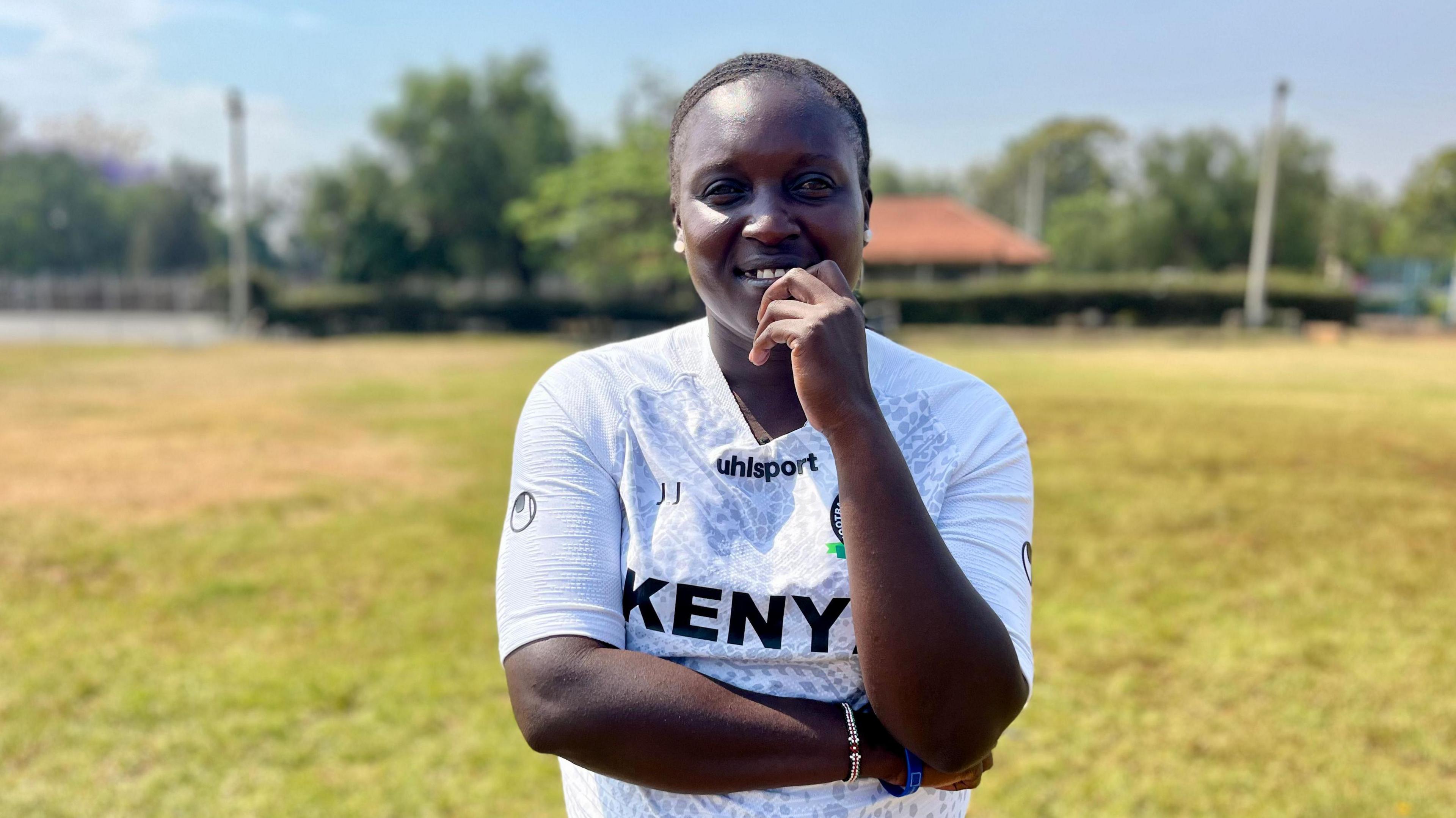 Jackline Juma, wearing a white training shirt with the word Kenya on it, smiles as she holds her left hand up towards her chin