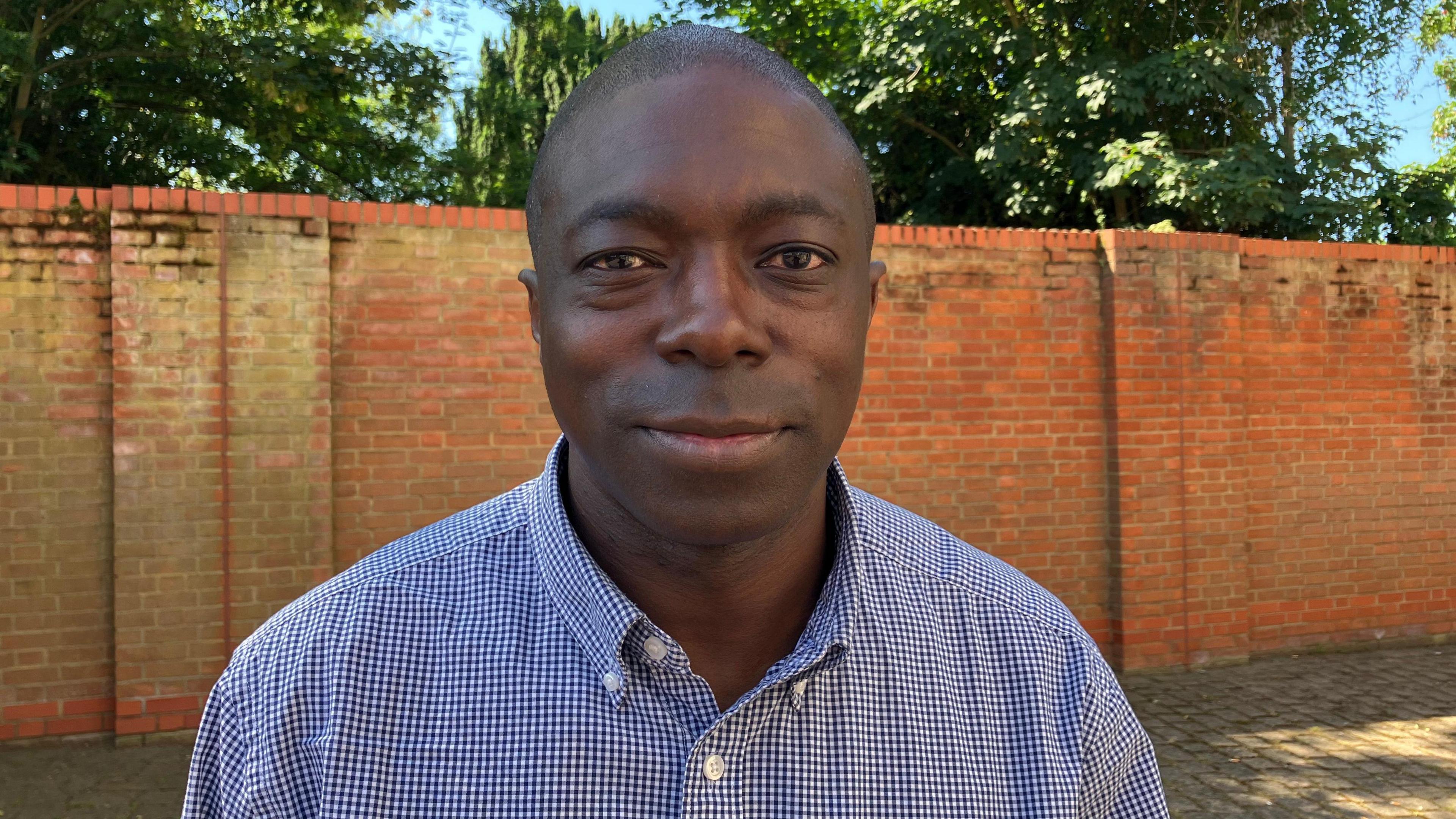 Bayo Alaba, standing in front of a brick wall with trees behind it. He wears a blue and white checked shirt with button-down collar.