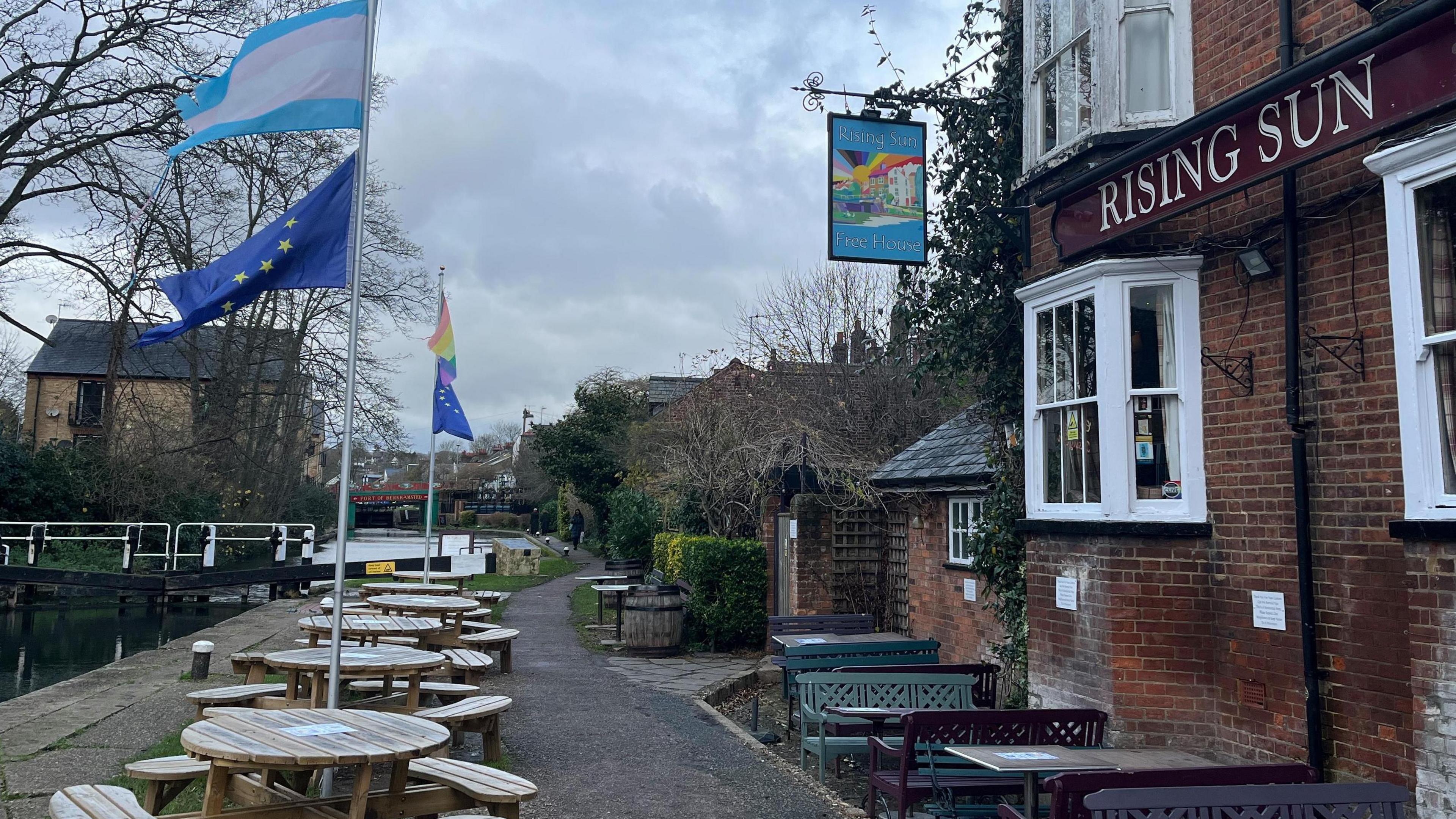 Outside of a pub with tables and benches and the canal running alongside. There is a lock in the background with a bridge beyond that.