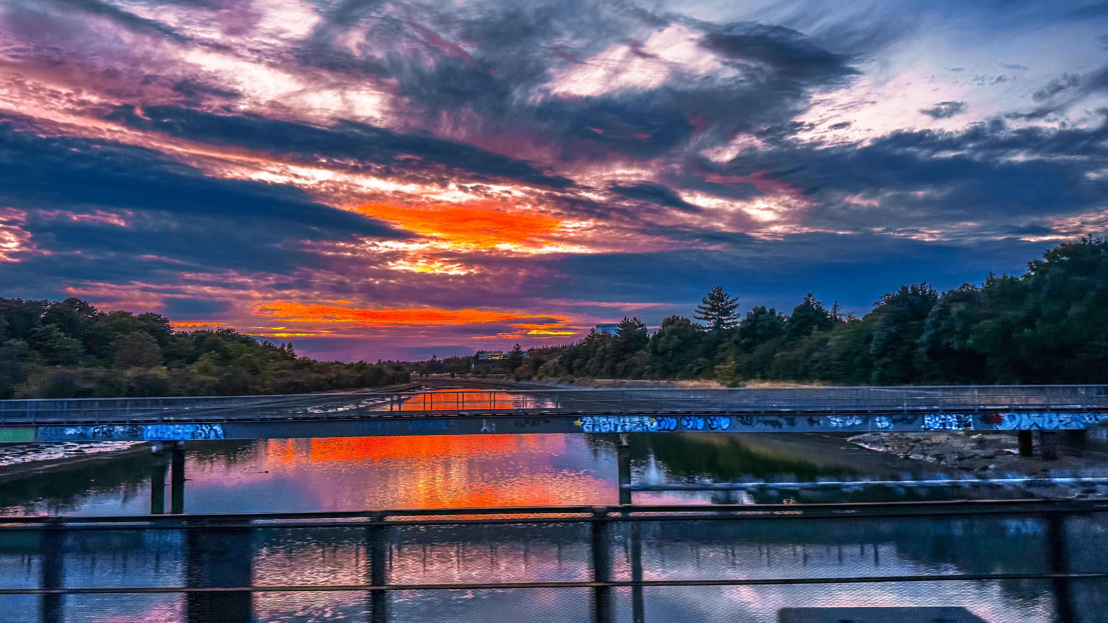 A bright orange sunset lights up the sky with vivid colours. The light is reflected in the water below making it glow a bright orange colour. The water has a grey bridge over it and the banks are flanked by trees.