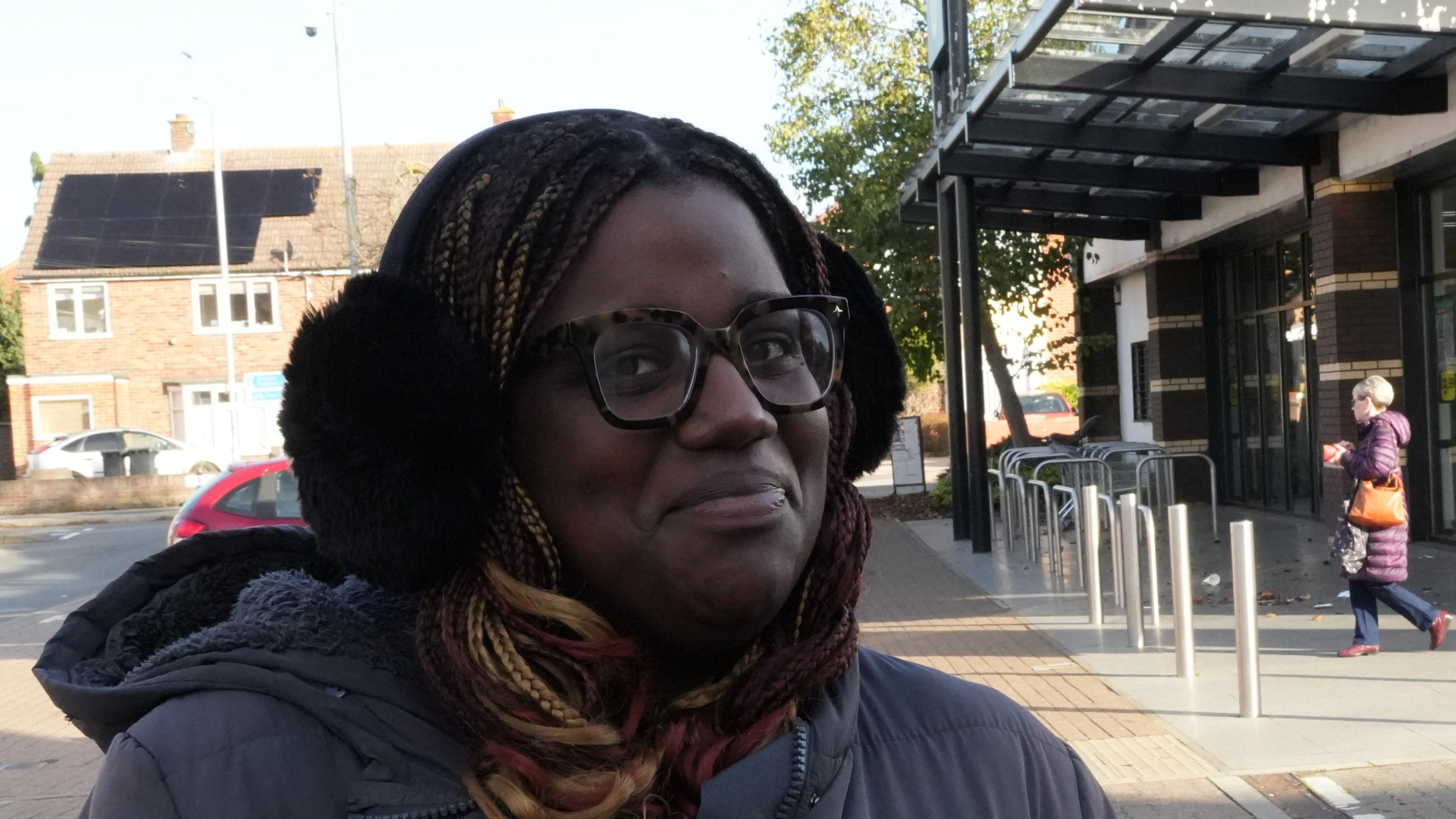Tia Bell smiles at the camera as she stands outside a supermarket. She wears black ear muffs, glasses and wears a dark blue coat. Some shoppers can be seen leaving the building behind her.