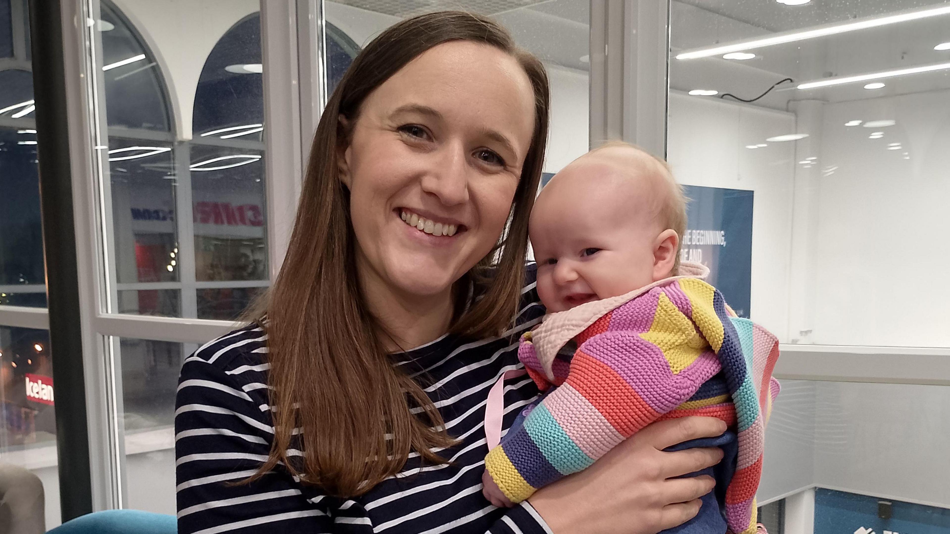 Christelle Lees is smiling as she holds a baby. The baby is smiling and wearing mustard coloured tights and a colourful cardigan.