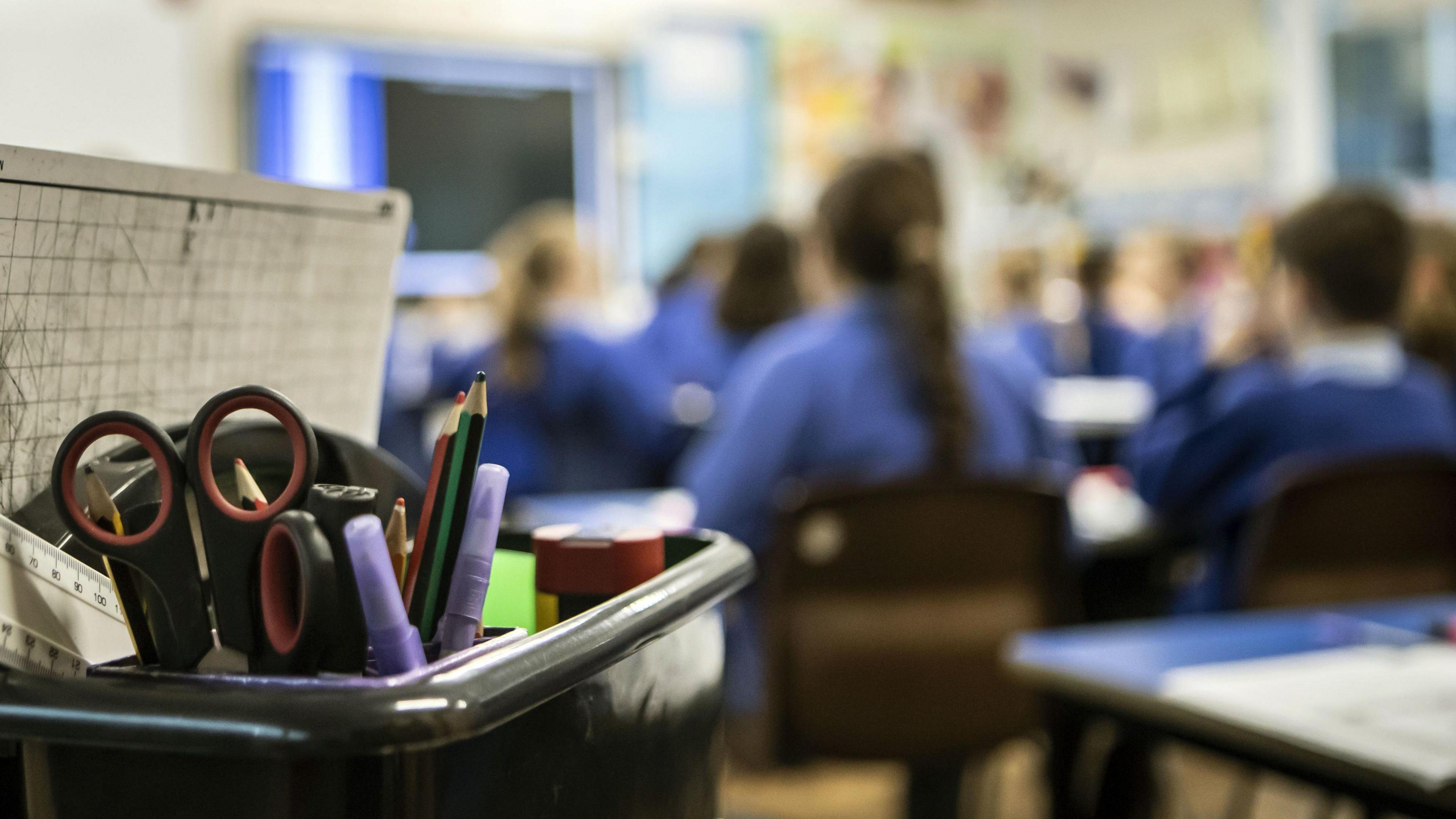 School children during a Year 5 class at a primary school, blurred background but in the foreground is a pencil pot with scissors, a ruler, pencils and highlighters in.