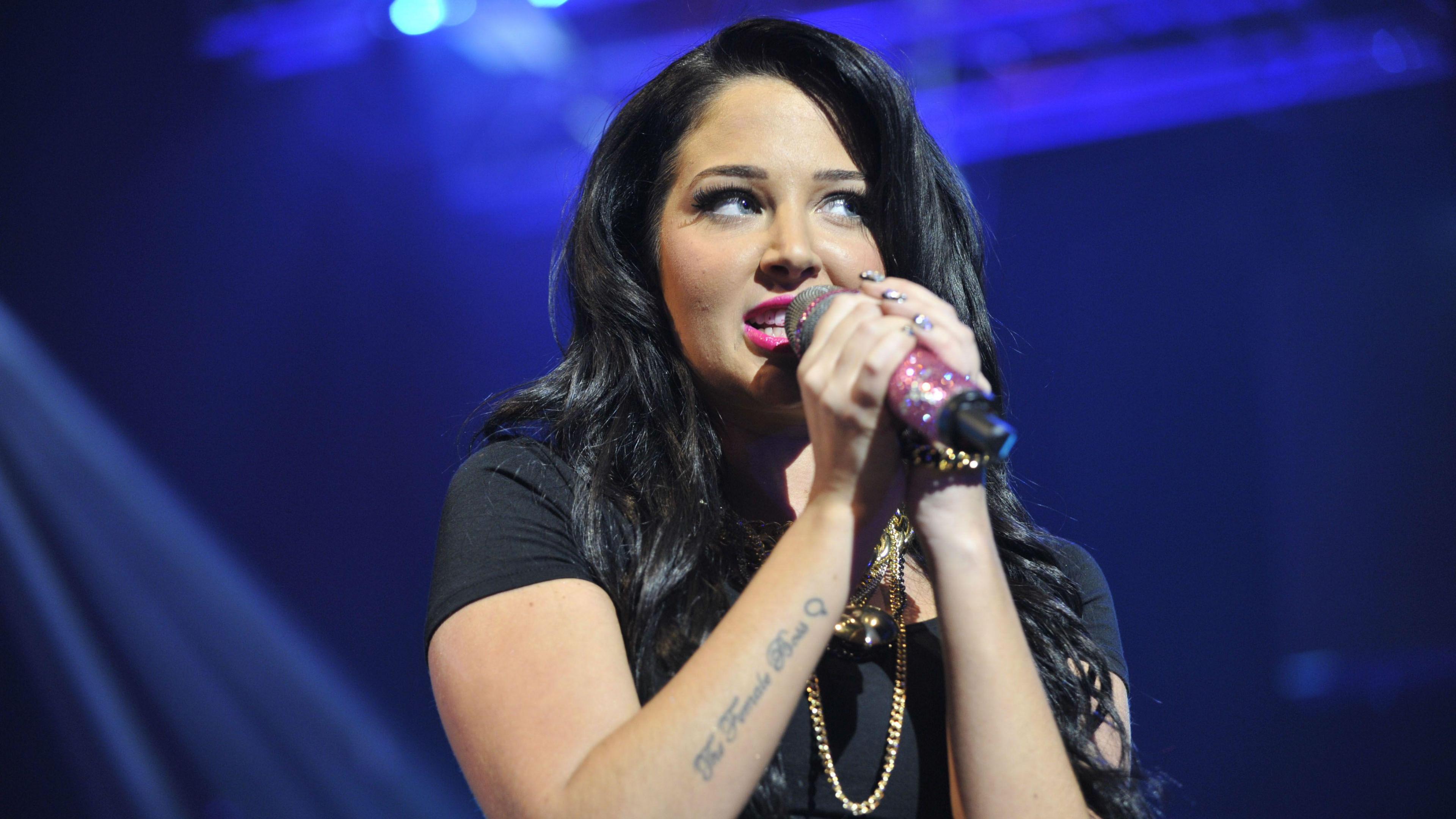 Tulisa singing into a microphone. She is wearing a black t-shirt and has hot pink lipstick on.