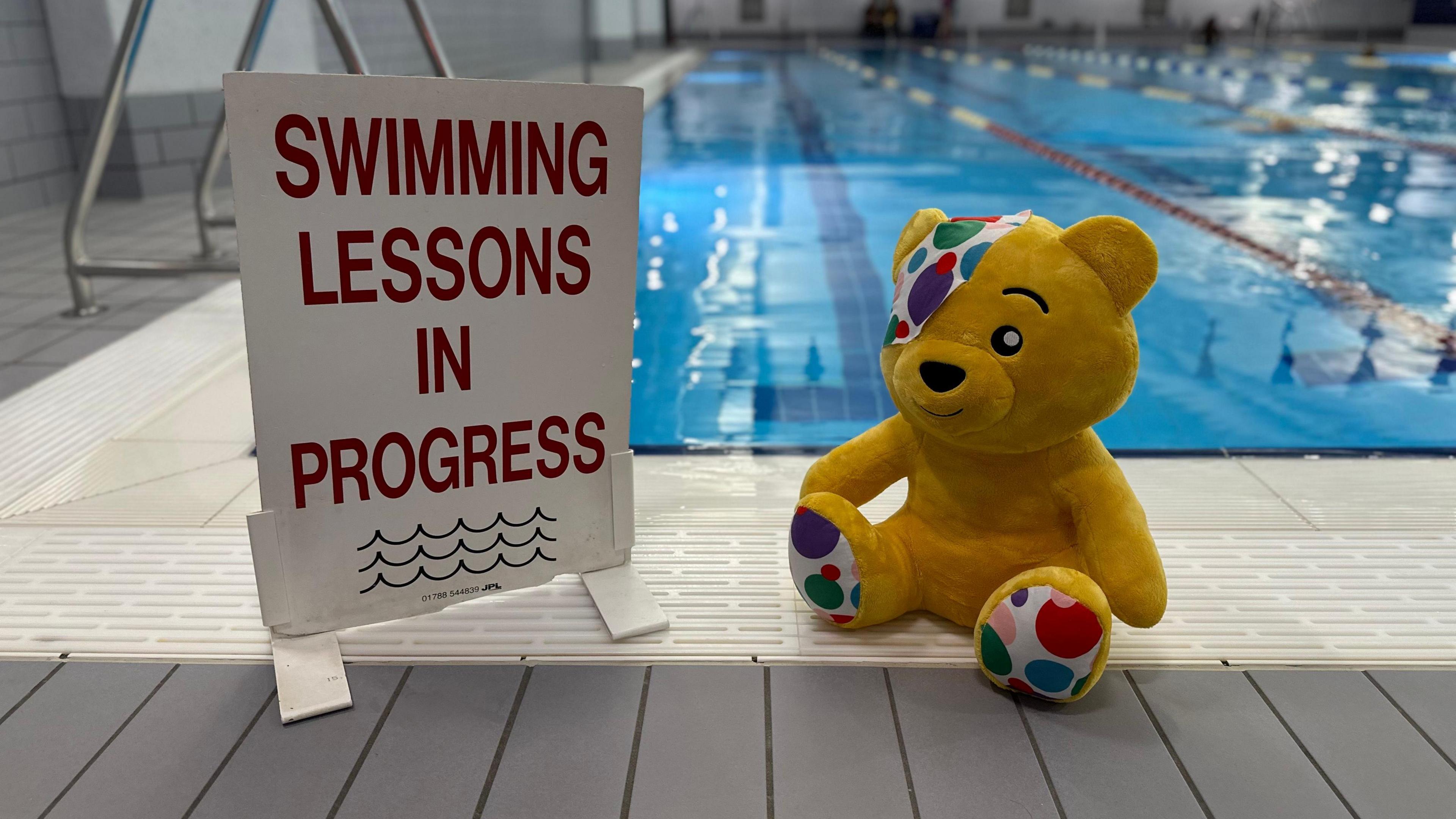 A sign says swimming lessons in proccess with some waves drawn on under the text. A toy Pudsey bear is sat next to it and behind the toy is a pool with some swimmers going up and down the lanes. Pudsey is a yellow bear with polka dots on the bottom of his feet and on his eye patch.