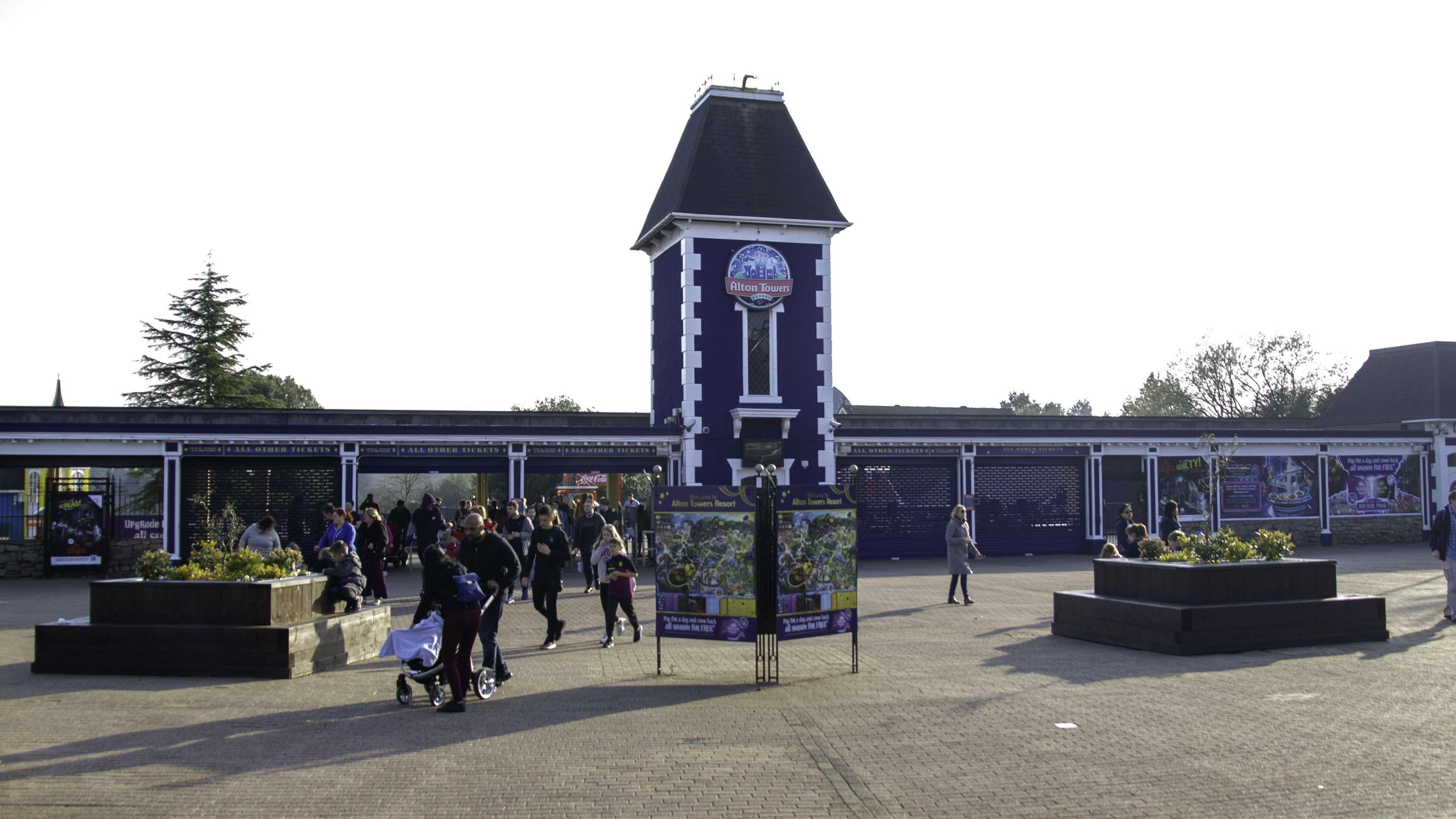 The entrance to Alton Towers theme park in Staffordshire. There is a tall central tower with the theme park's logo on it. There are people milling about on the concourse in front of the entrace.