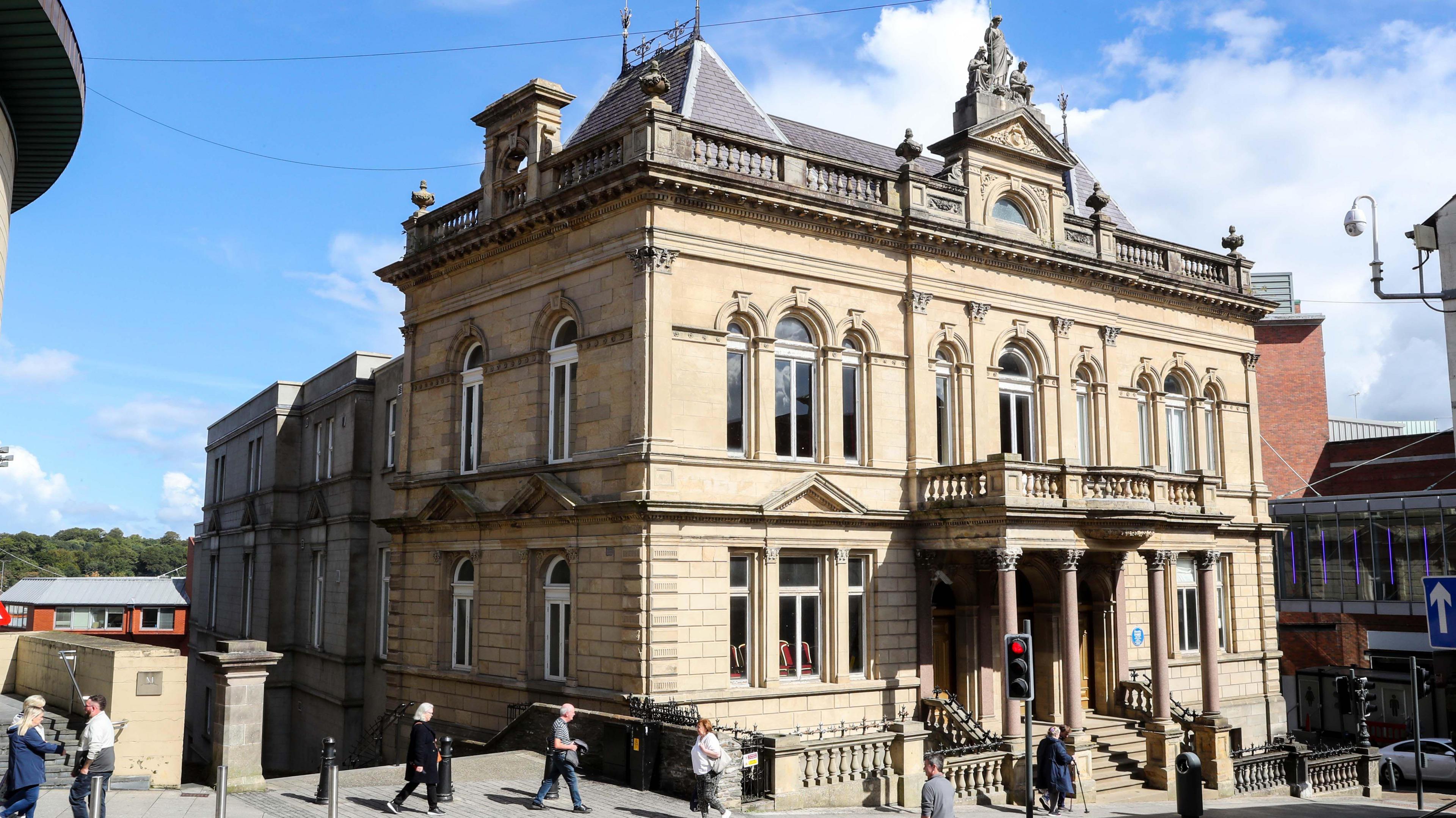 A grand old hall stands on a busy street in Londonderry, it has five storeys and a balcony can be seen to the front of the building, supported by columns. A number of pedestrians pass by.