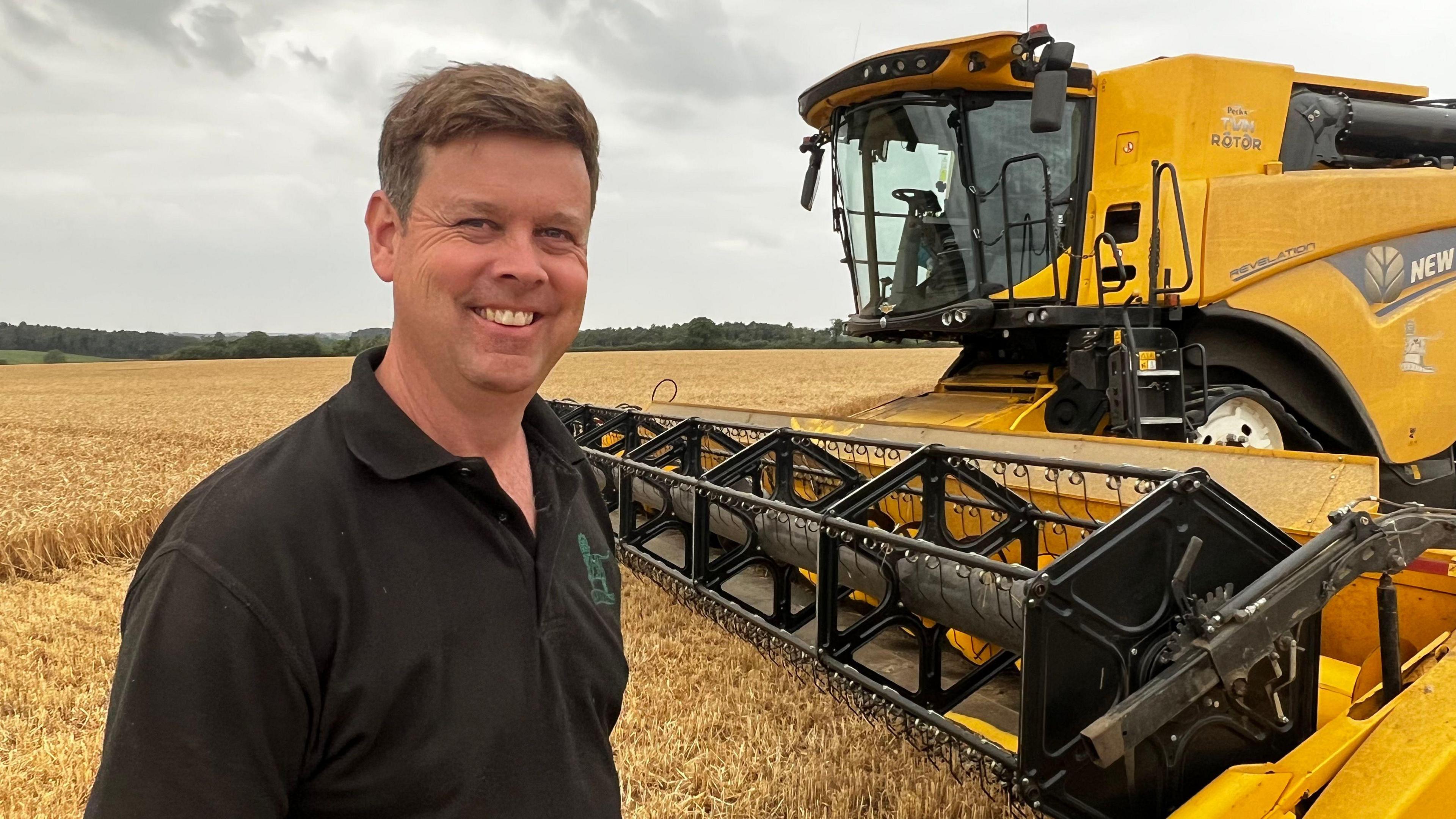 Robin Aird smiles at the camera, wearing a black polo top. he stands in a golden wheat field, with the huge yellow combine harvester to the right.