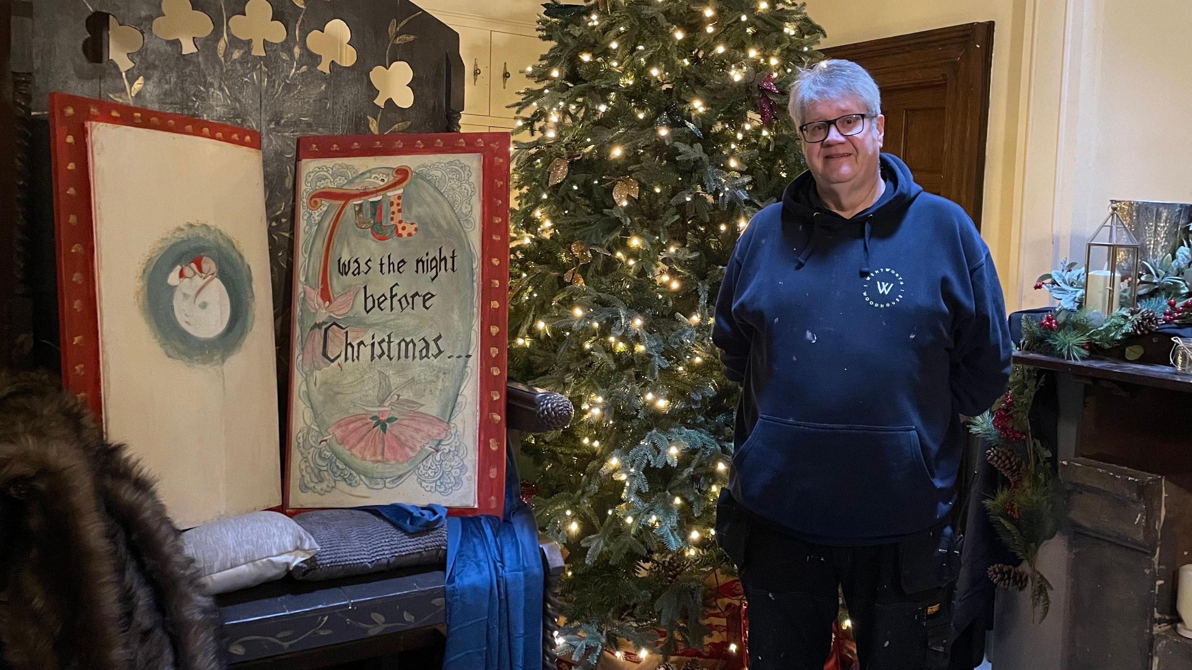 Neil is stood in a hoodie covered in specks of paint. Behind him is a lit and decorated Christmas tree, and next to him is a wooden throne. On the throne is a large prop that looks like a book, with hand-painted pages. The book reads: "T'was the night before Christmas."