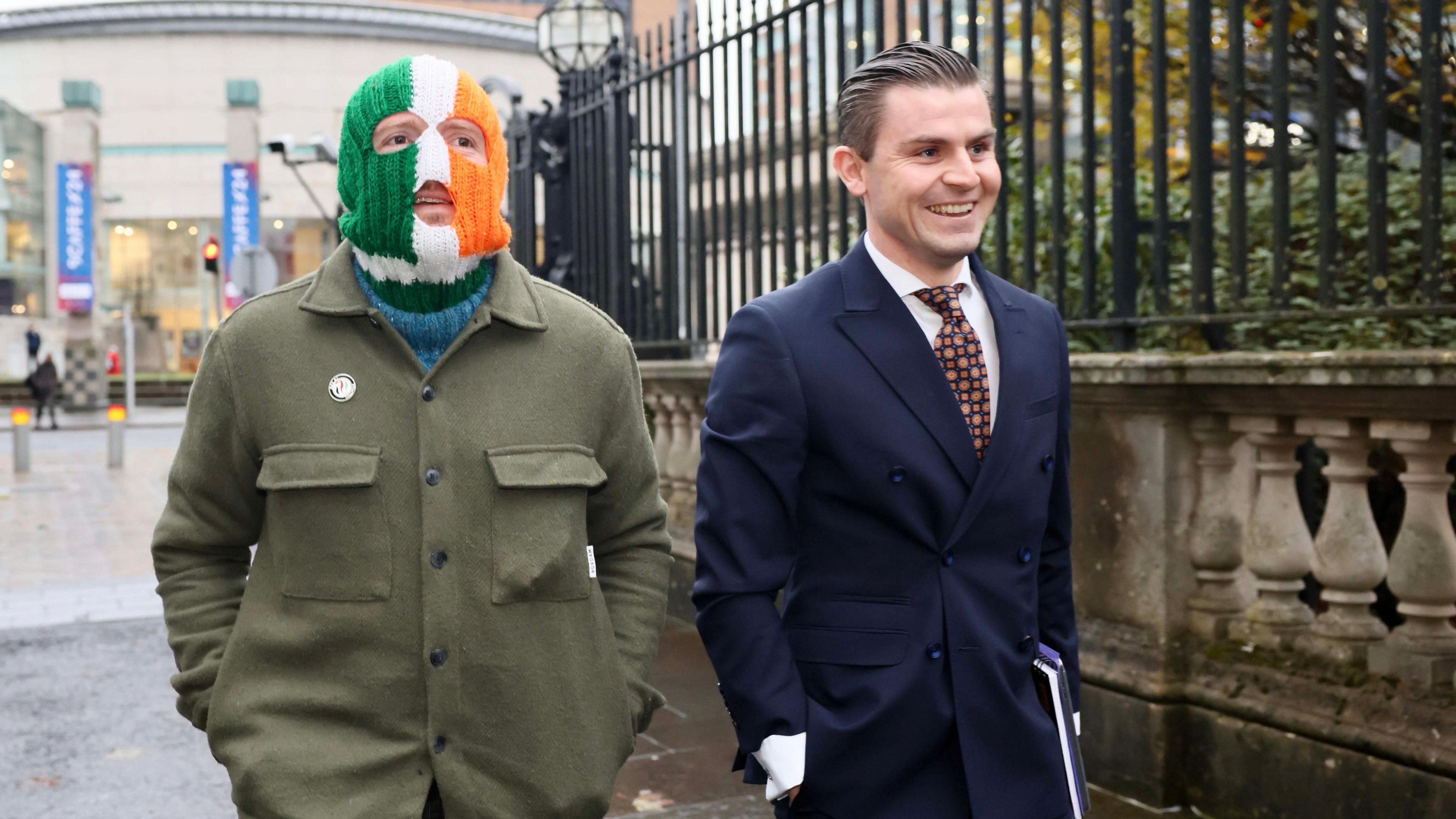 Two men talking along a street. One is wearing a green coast and a knitted balaclava in the colours of the Irish flag