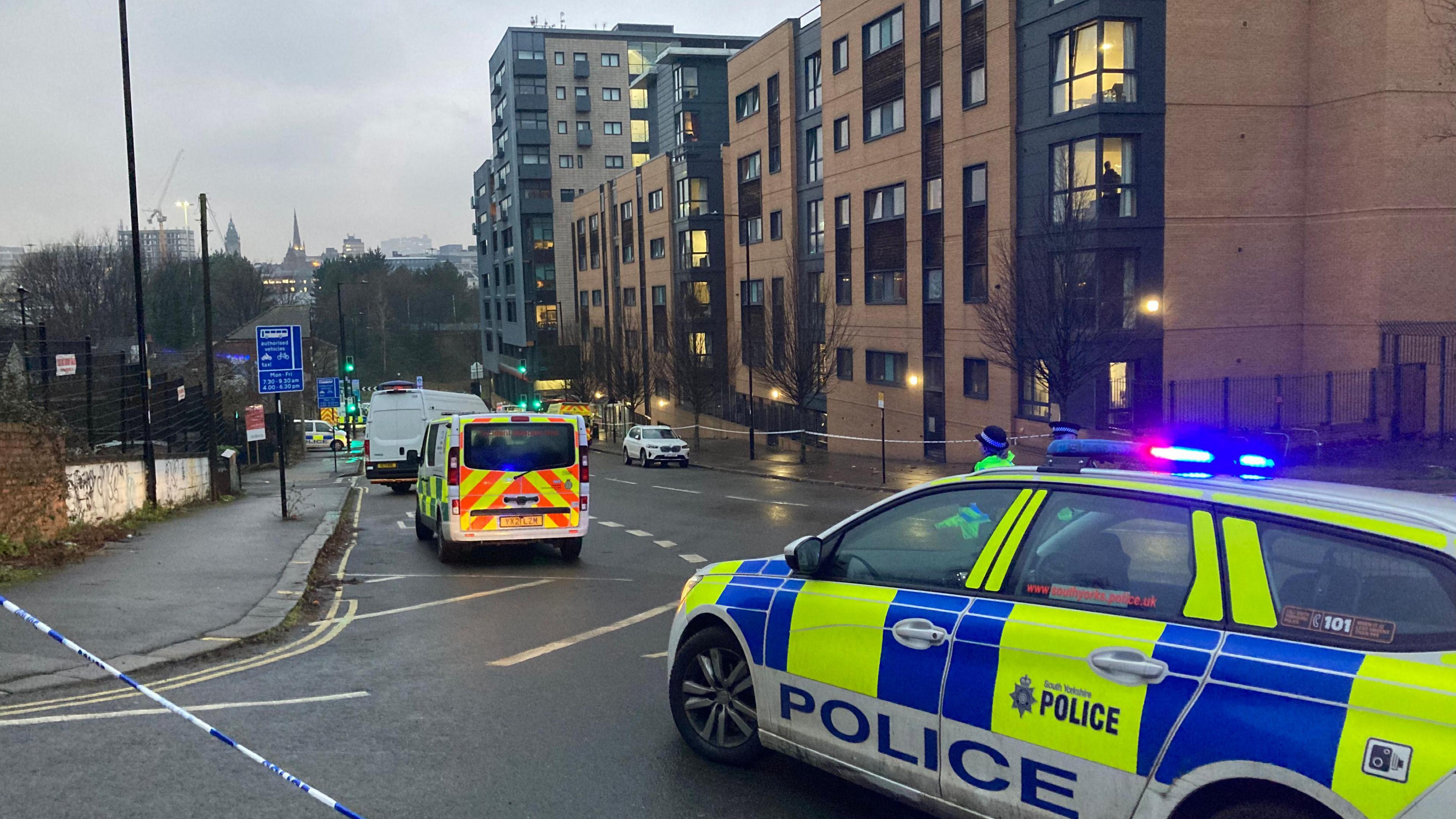 Police vehicles parked on the side of a road, next to a police cordon. There are blocks of flats on the other side of the road. The weather looks gloomy and grey.