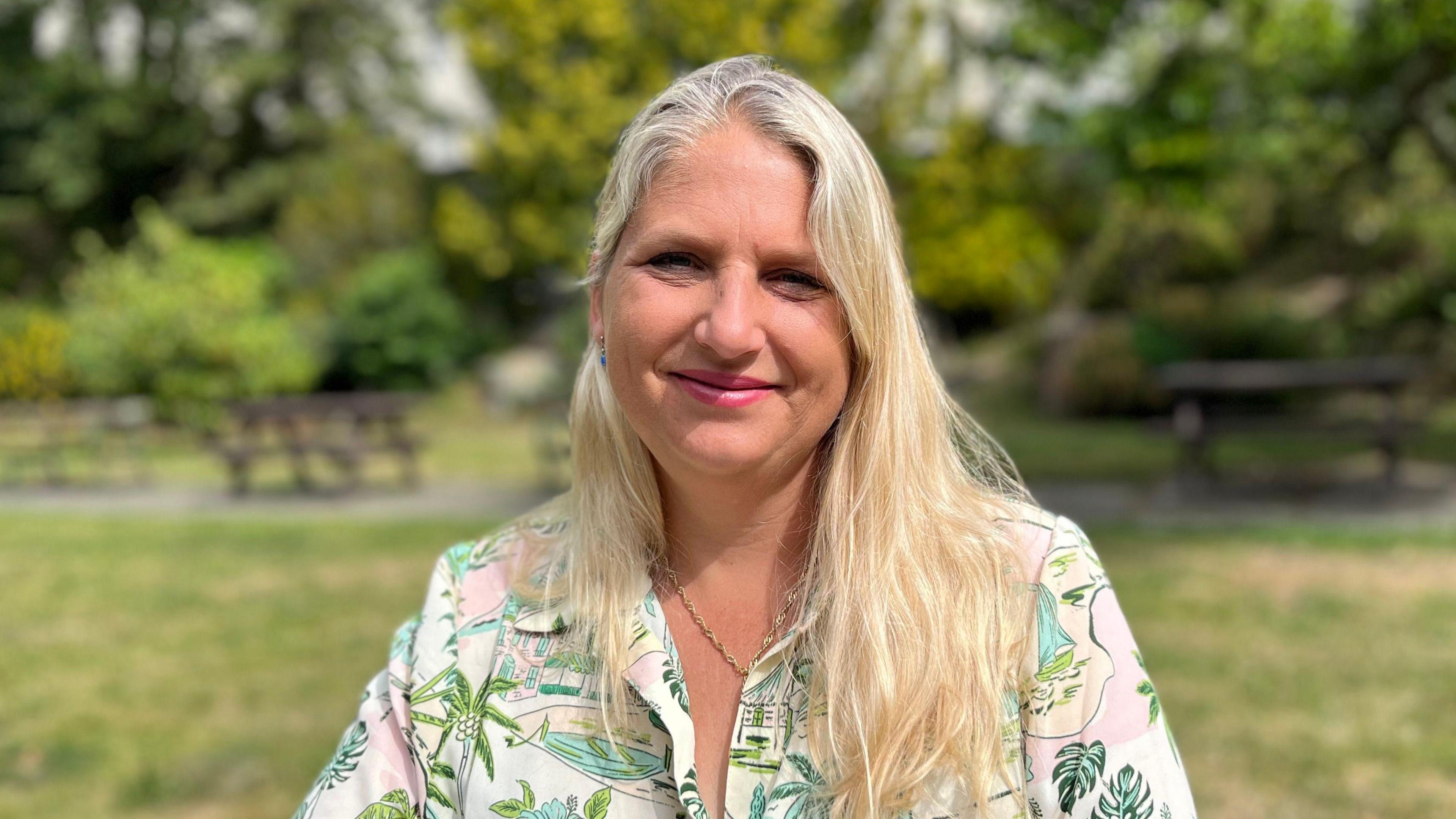 Victoria Oliver smiling at the camera. She is sitting in a park area with picnic benches, grass and trees in the background.