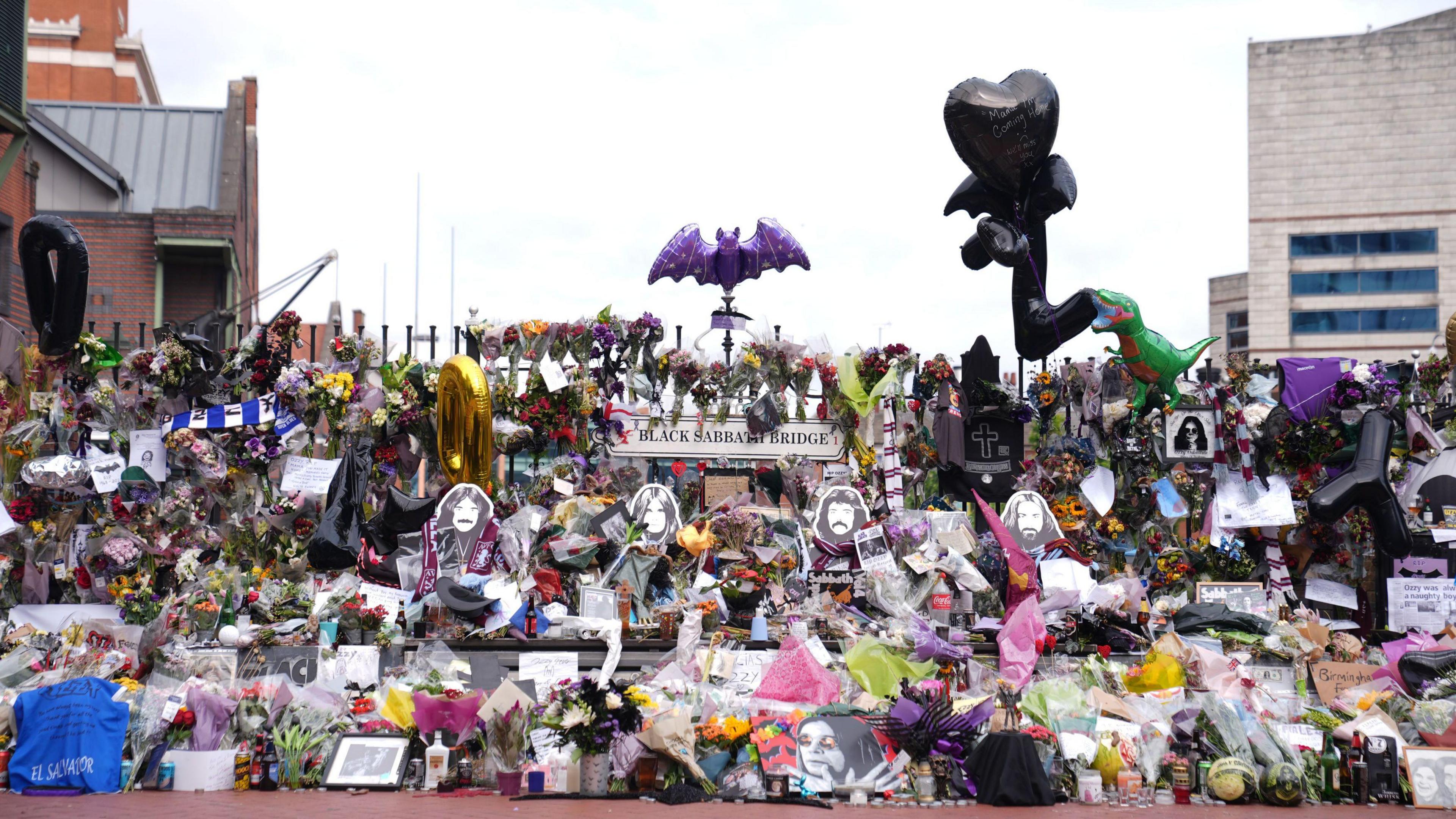 A picture from the front of the Black Sabbath bench, which has cutouts of the four black sabbath members on it. On and around the bench are thousands of bunches of flowers, cards, and photos of Ozzy Osbourne. There is a large purple bat balloon tied to a fence, as well as a black bat balloon, and a black heart balloon.