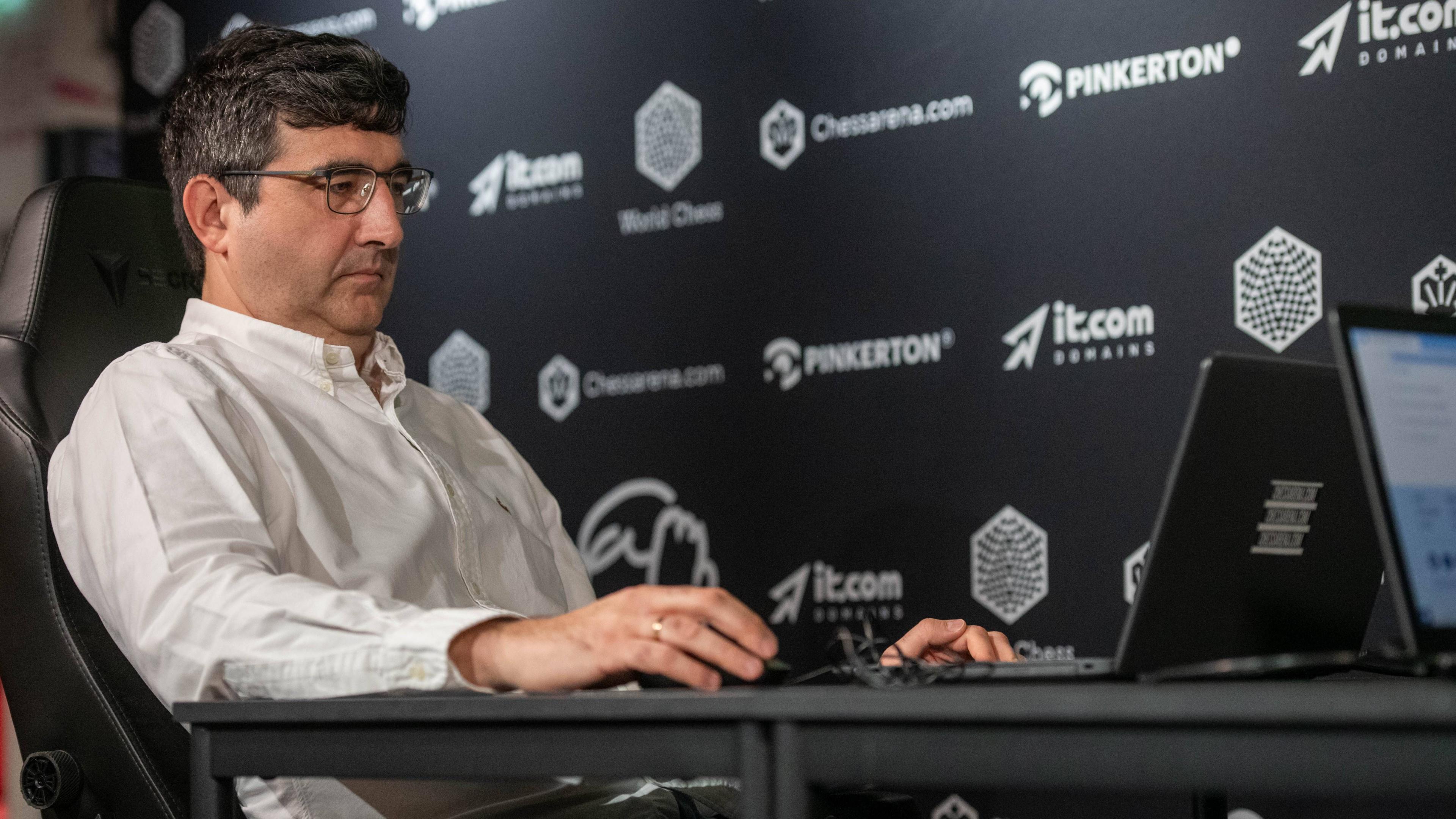Vladimir Kramnik at a chess tournament in London in August 2024. He sits facing his opponent and is wearing a white shirt and glasses.