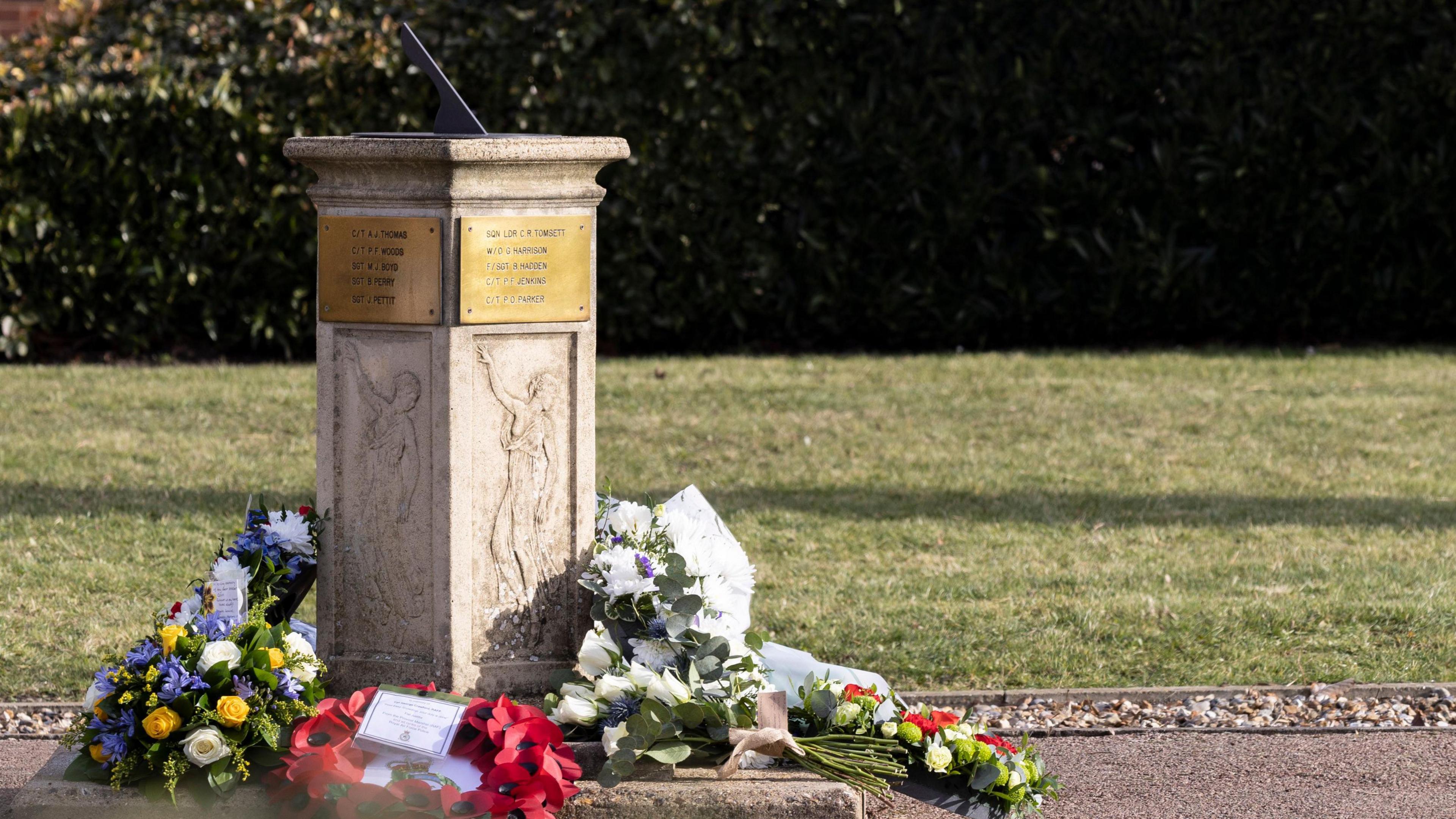 A stone sundial has flowers, including a wreathe of poppies, laid next to it. Gold plaques on each side of the sundial lists names of people killed in the bus crash.