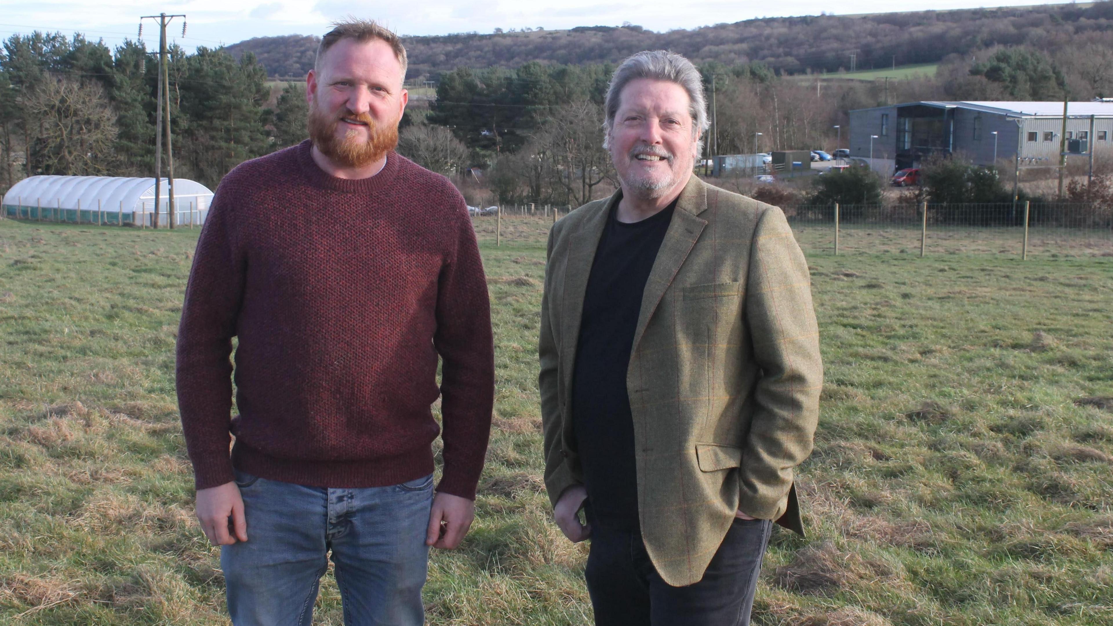 Dave Wilde, who has a ginger beard, is wearing a claret jumper and blue jeans. John Phillipson is wearing a green/brown blazer and a black round neck top with dark trousers. They are standing next to each other in field.