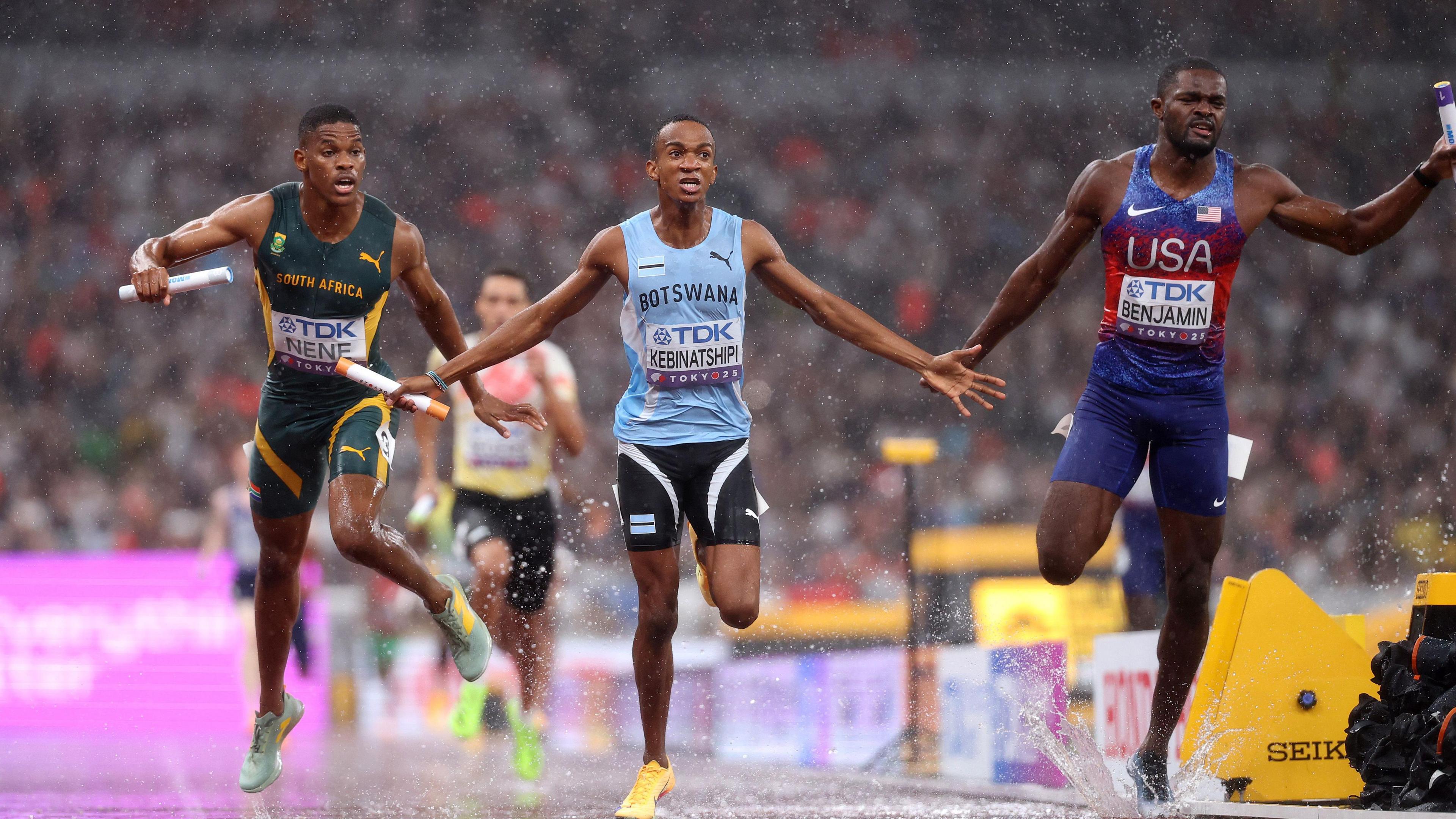 Collen Kebinatshipi in the light blue of Botswana (centre) stretches his arms out in celebration as he crosses the finish line to win the gold medal in the men's 4x400m relay at the World Athletics Championships in Tokyo. Zakithi Nene of South Africa is seen to the left and Rai Benjamin of the United States is on the right. All are holding batons and causing water to splash up from the track as they race in the rain