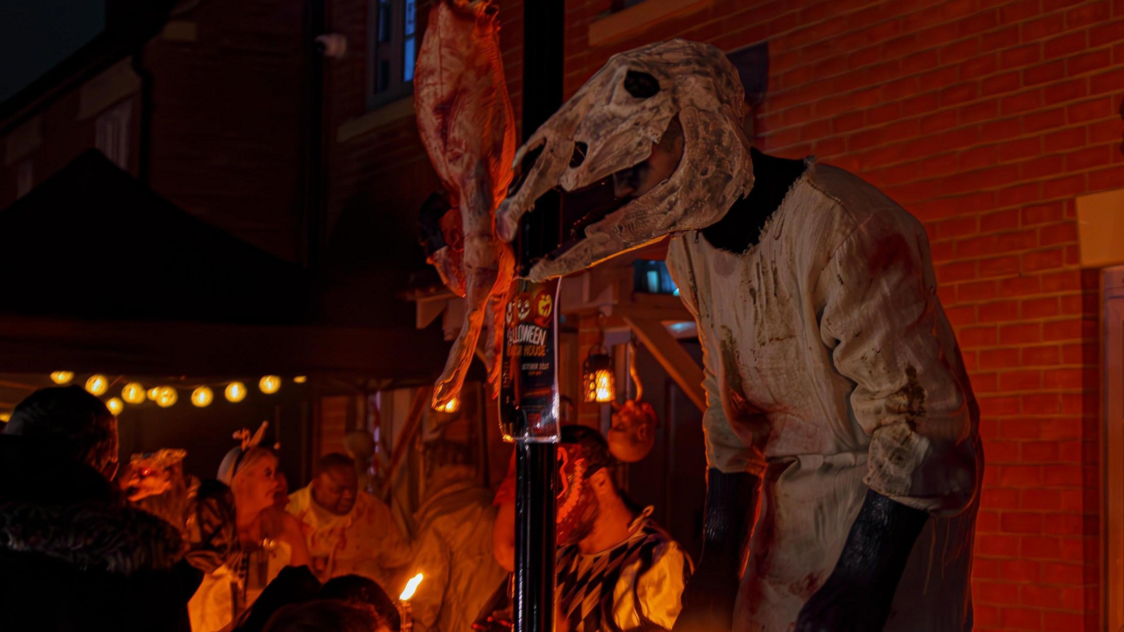 A person wearing a horse head skeleton costume and dressed in a bloody white robe stands beside the entrance to the Brackley Horror House 