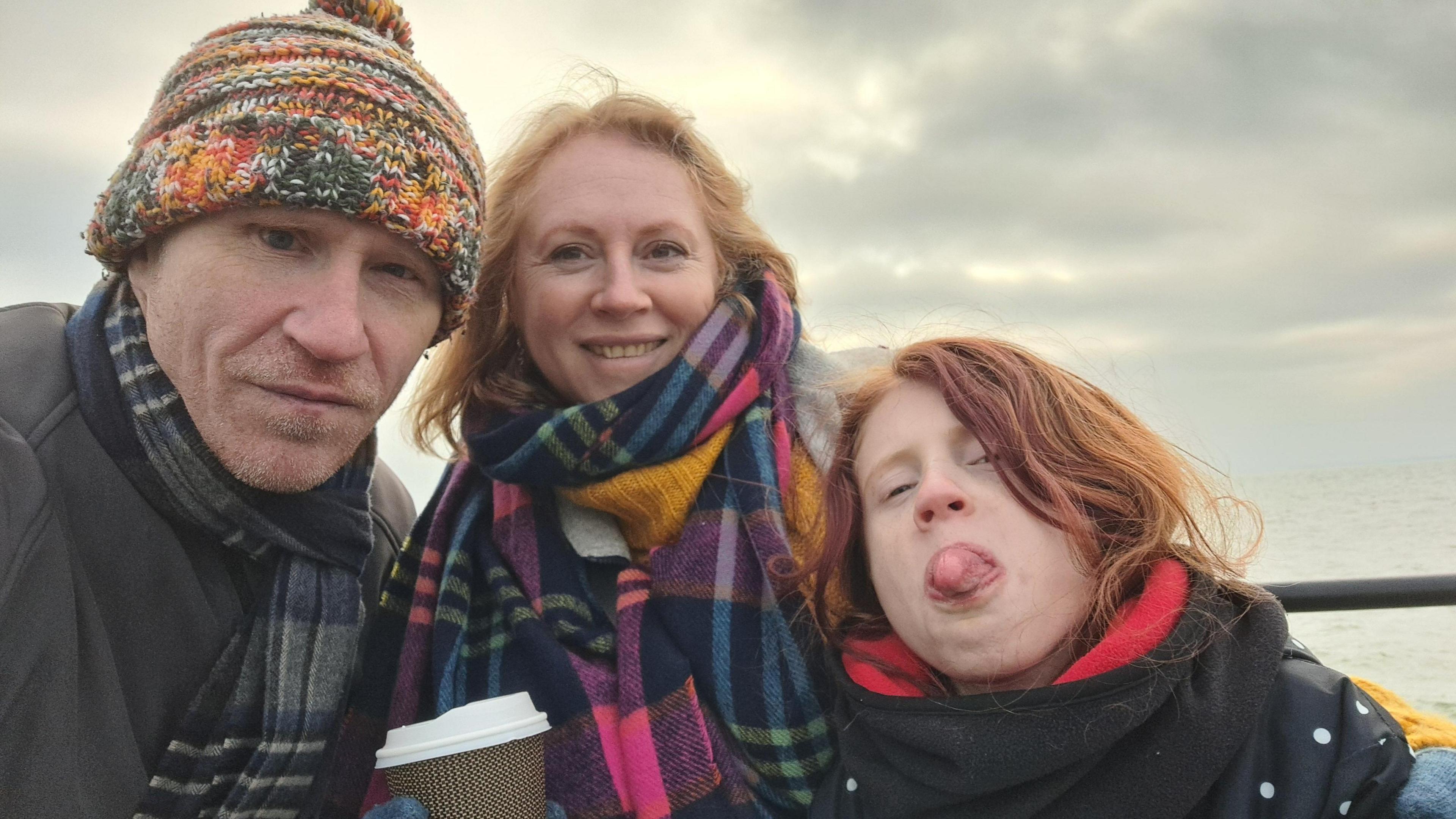 Paul Jefferies is standing next to his wife Fay and daughter Eirlys. In the background is the sea. They are wrapped up in scarves and coats. Paul and Fay are smiling and Eirlys is poking her tongue out.