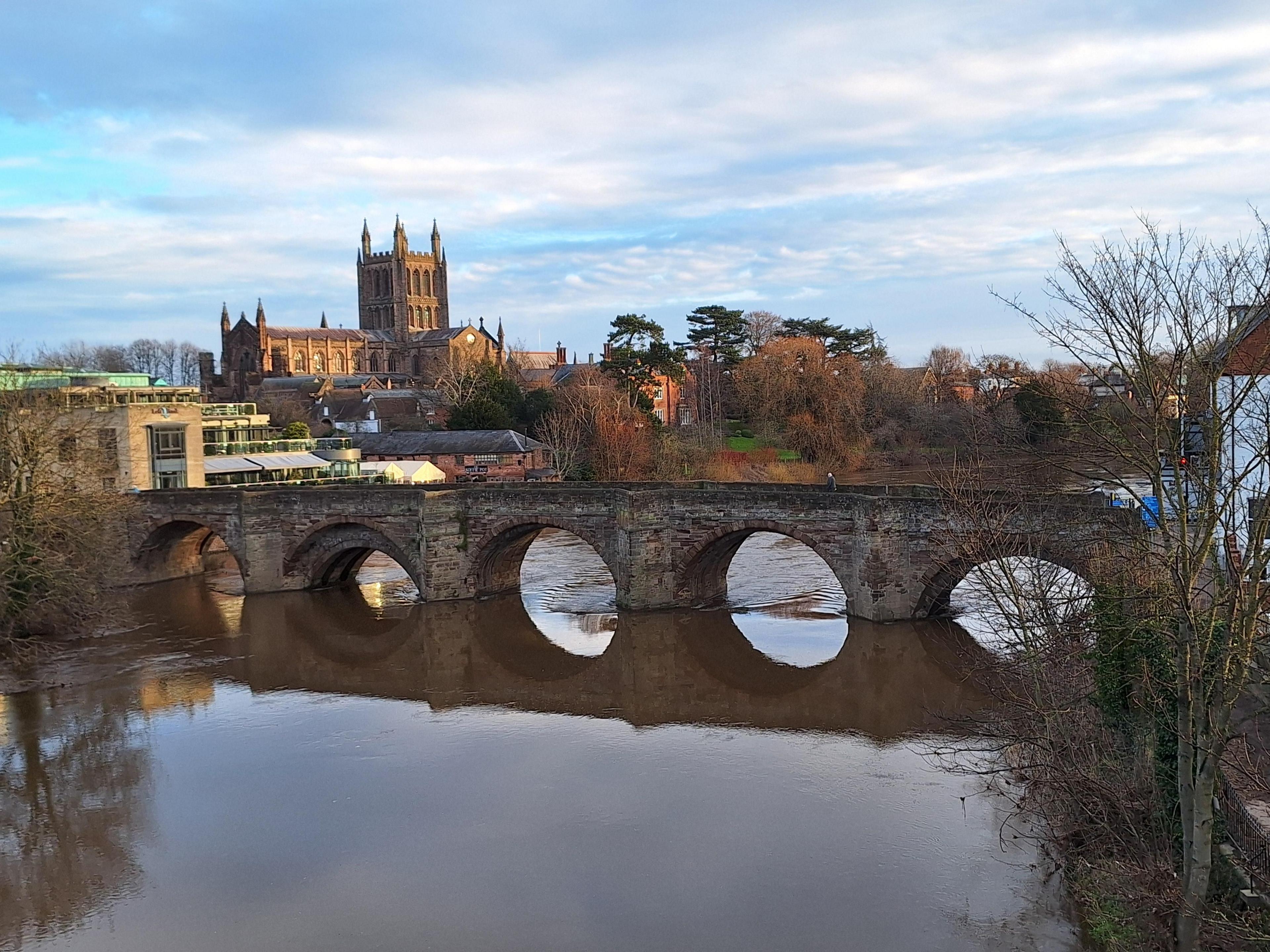 The river is in the foreground with a stone bridge across it in the middle of the shot and the cathedral slightly raised on a hillside behind. Bare trees encroach from the sides of the shot.