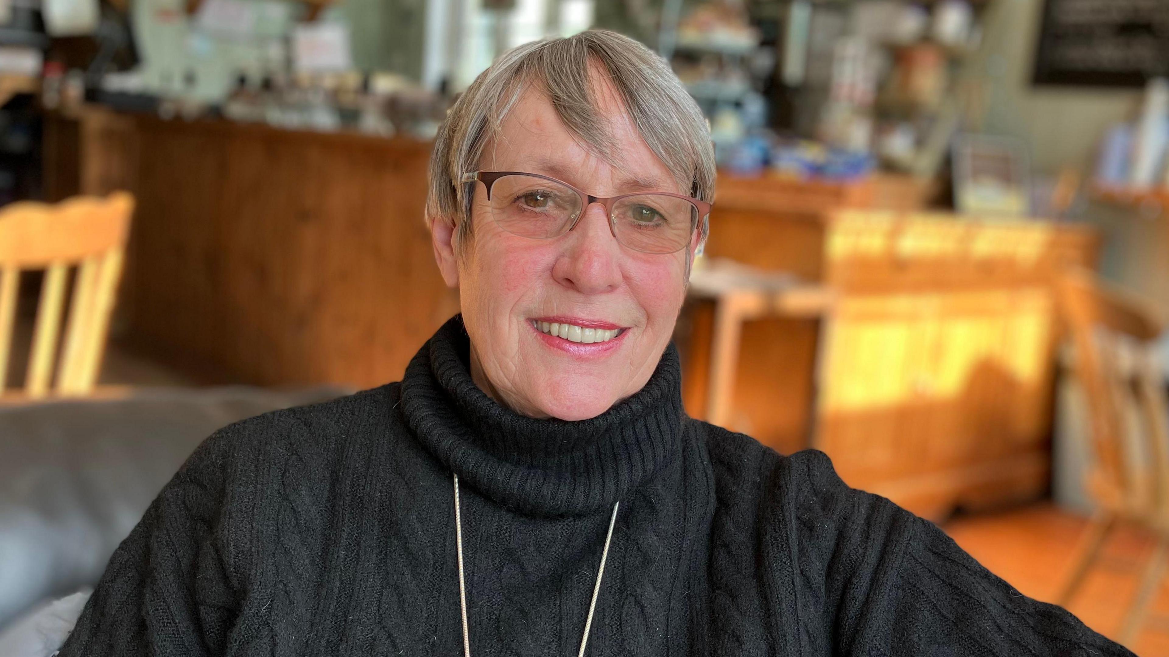 A woman with short, white hair. She is wearing a black turtle neck jumper with a necklace on top.