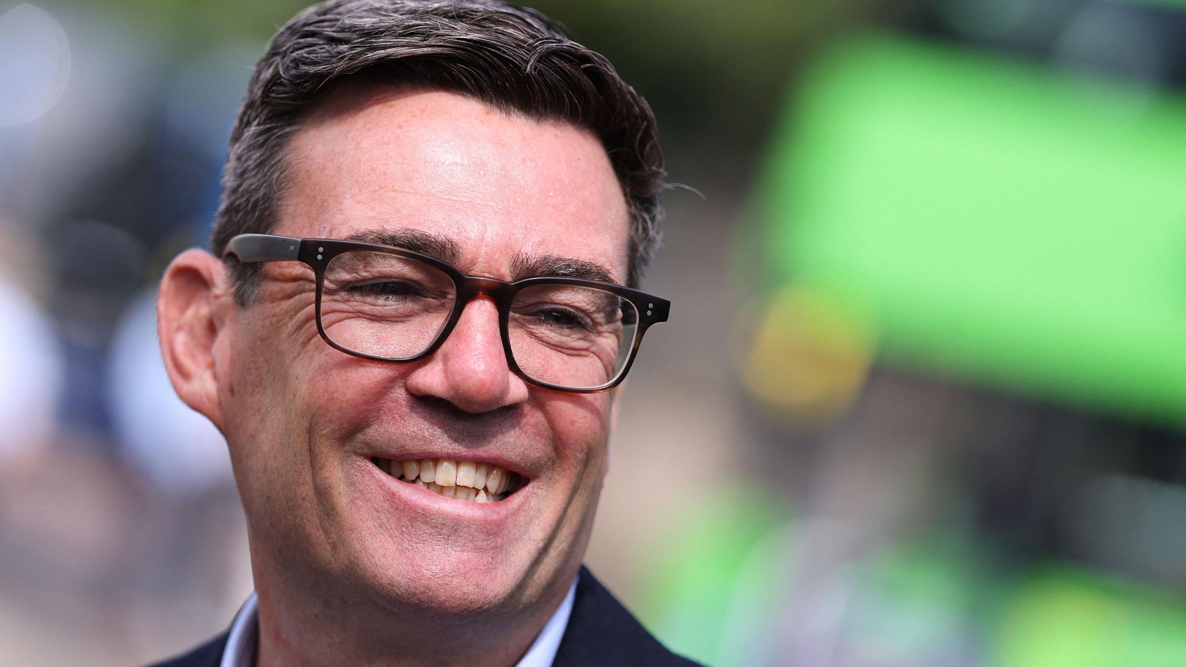 Andy Burnham is smiling and looking to the side. He has dark hair, brushed to one side and dark glasses.