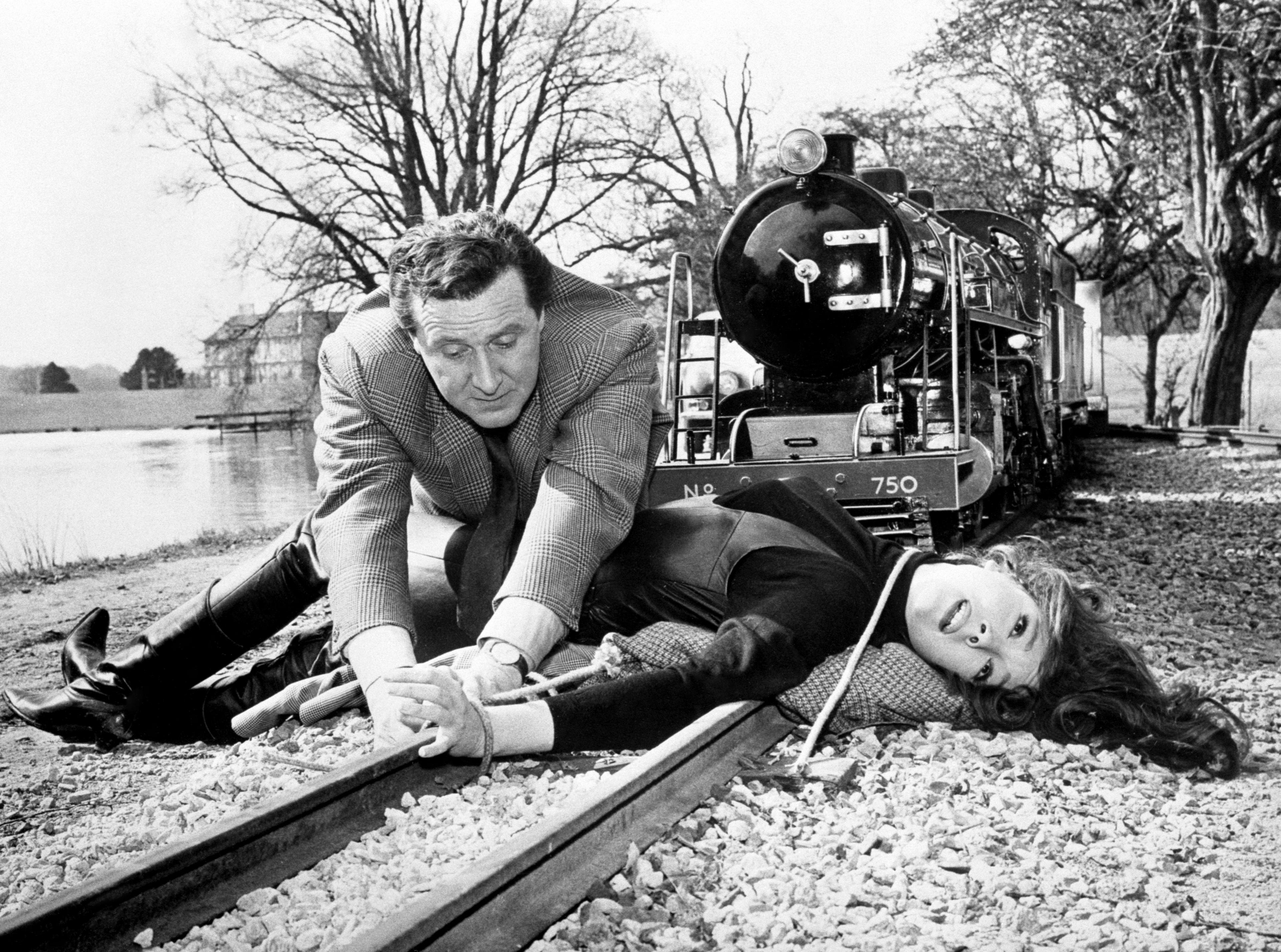 A black and white photo of a man untying ropes attached to a woman who is laid prone across a railway track, with a train posed just behind them