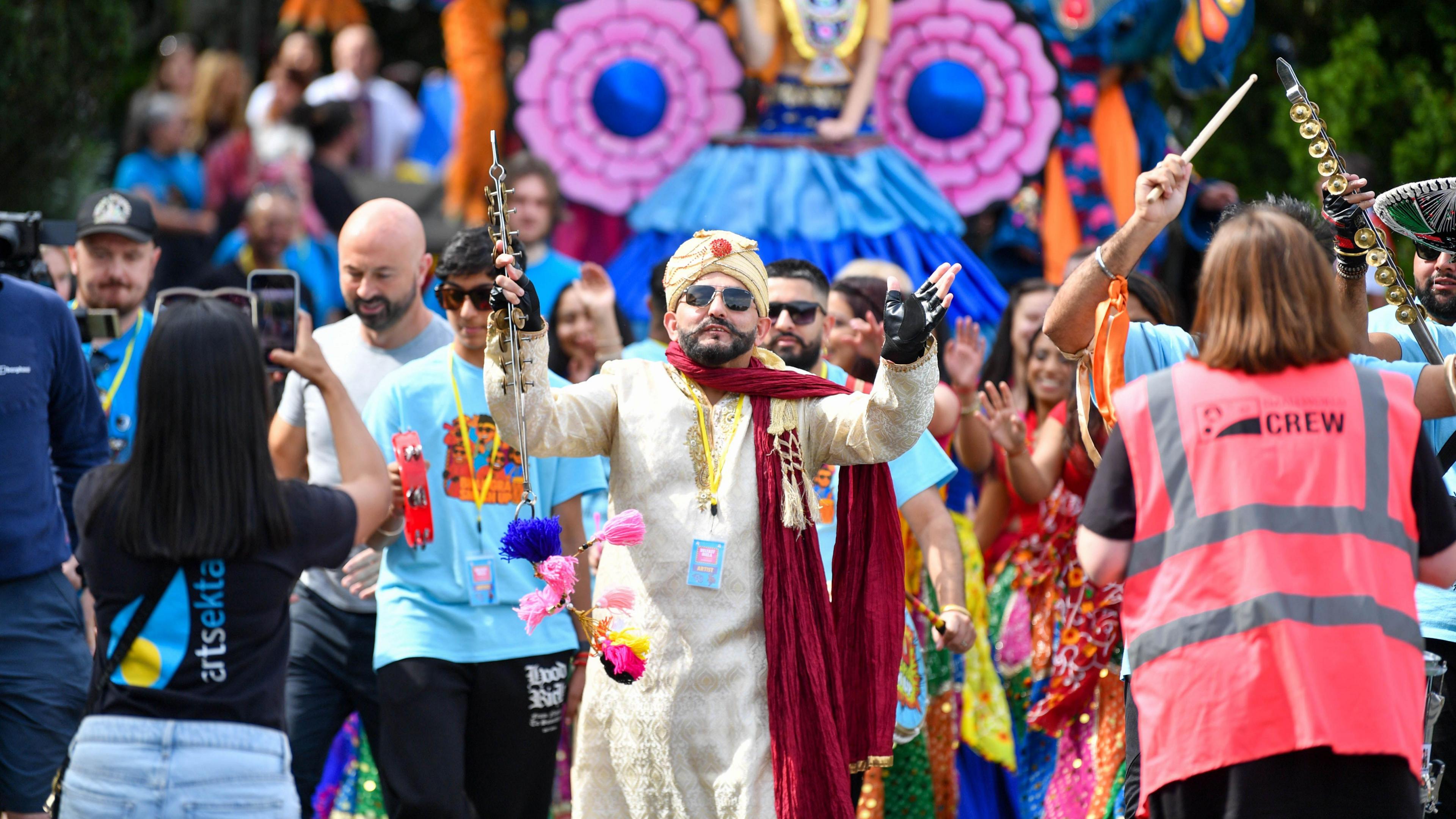 A man is taking part in a celebration. People dressed in various colours and costumes are in attendance. Crowds of people are standing around watching. 
