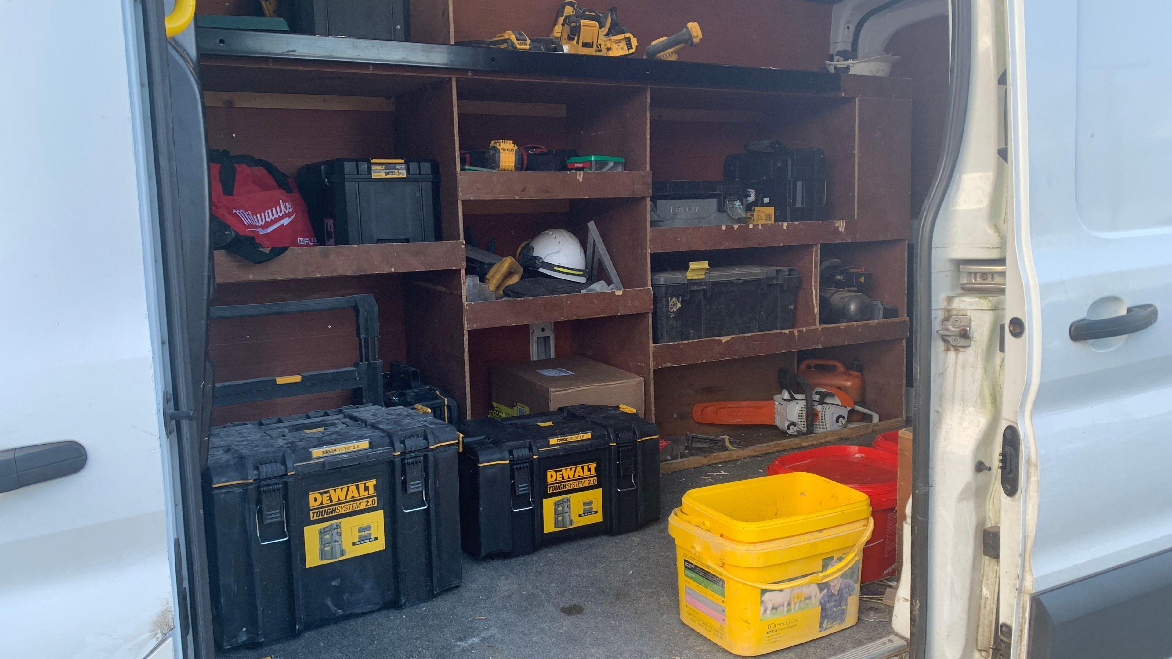 A side door of a white van is opened to show a wall full of tools. There are shelves filled with all kinds of power tools and boxes of tools.