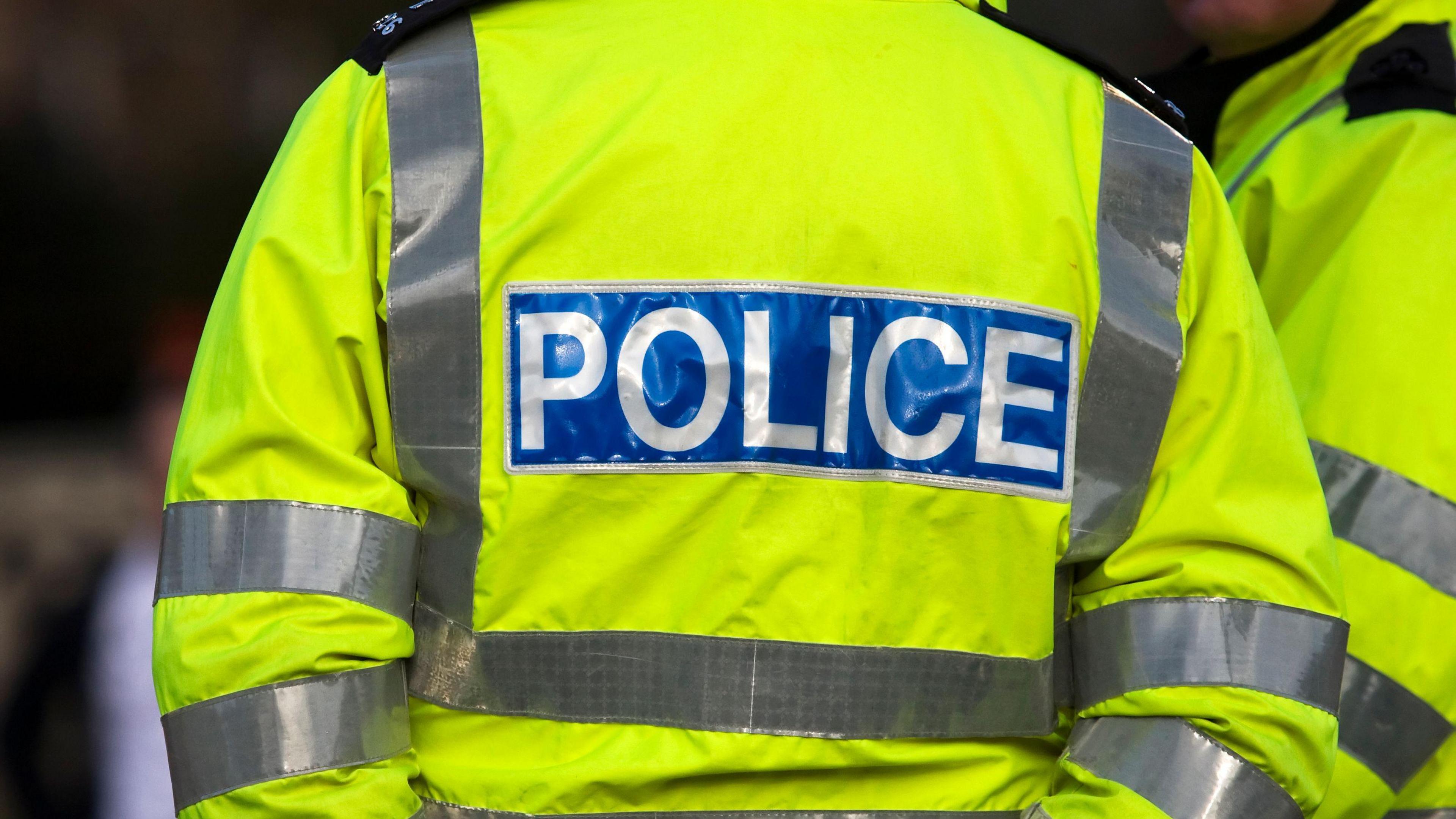 A police officer pictured from behind, wearing a hi-vis POLICE jacket and holding his hands behind his back