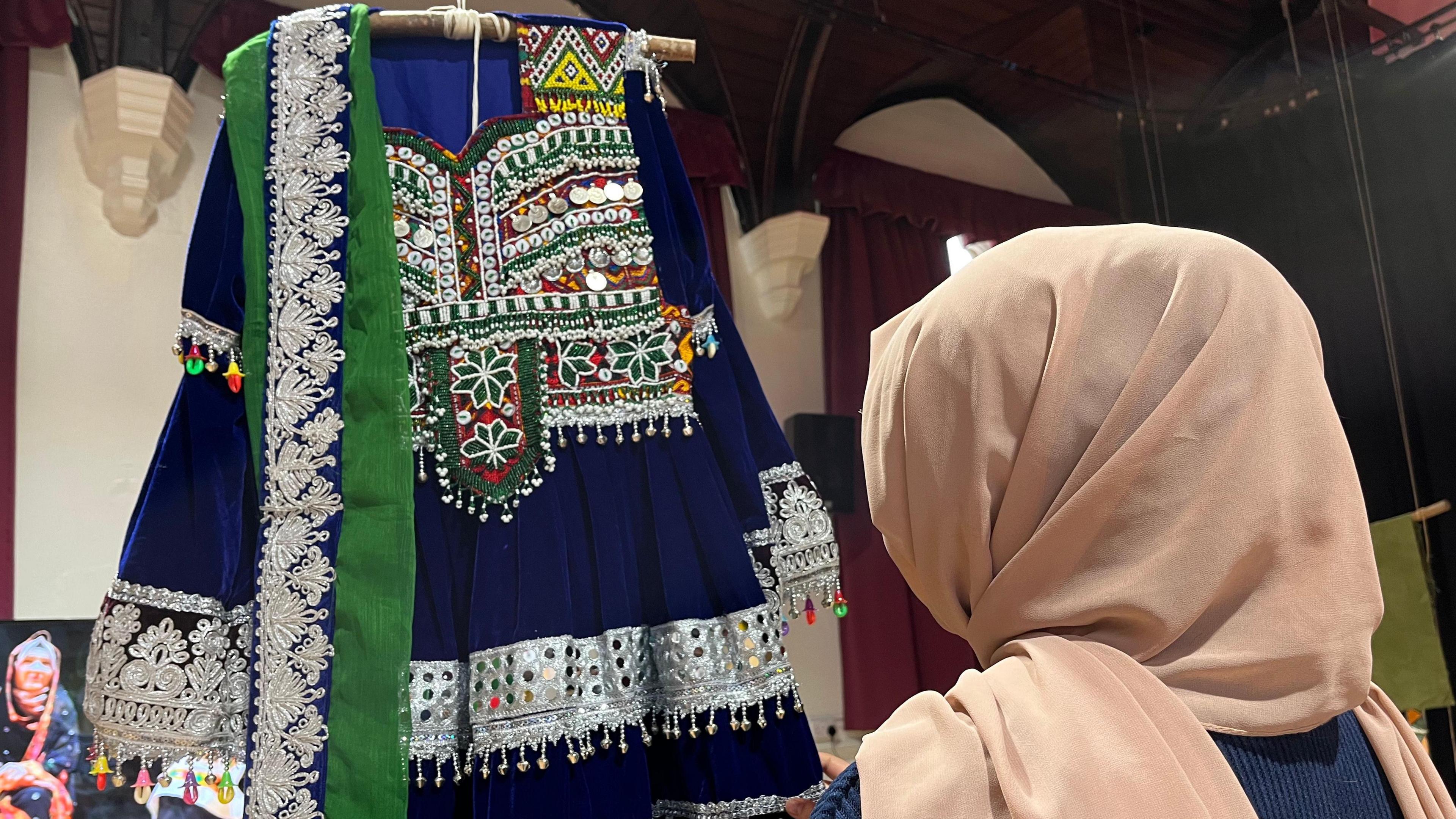 Afghan woman wearing a hijab stands infront of a traditional Afghan dress, we see her from behind 