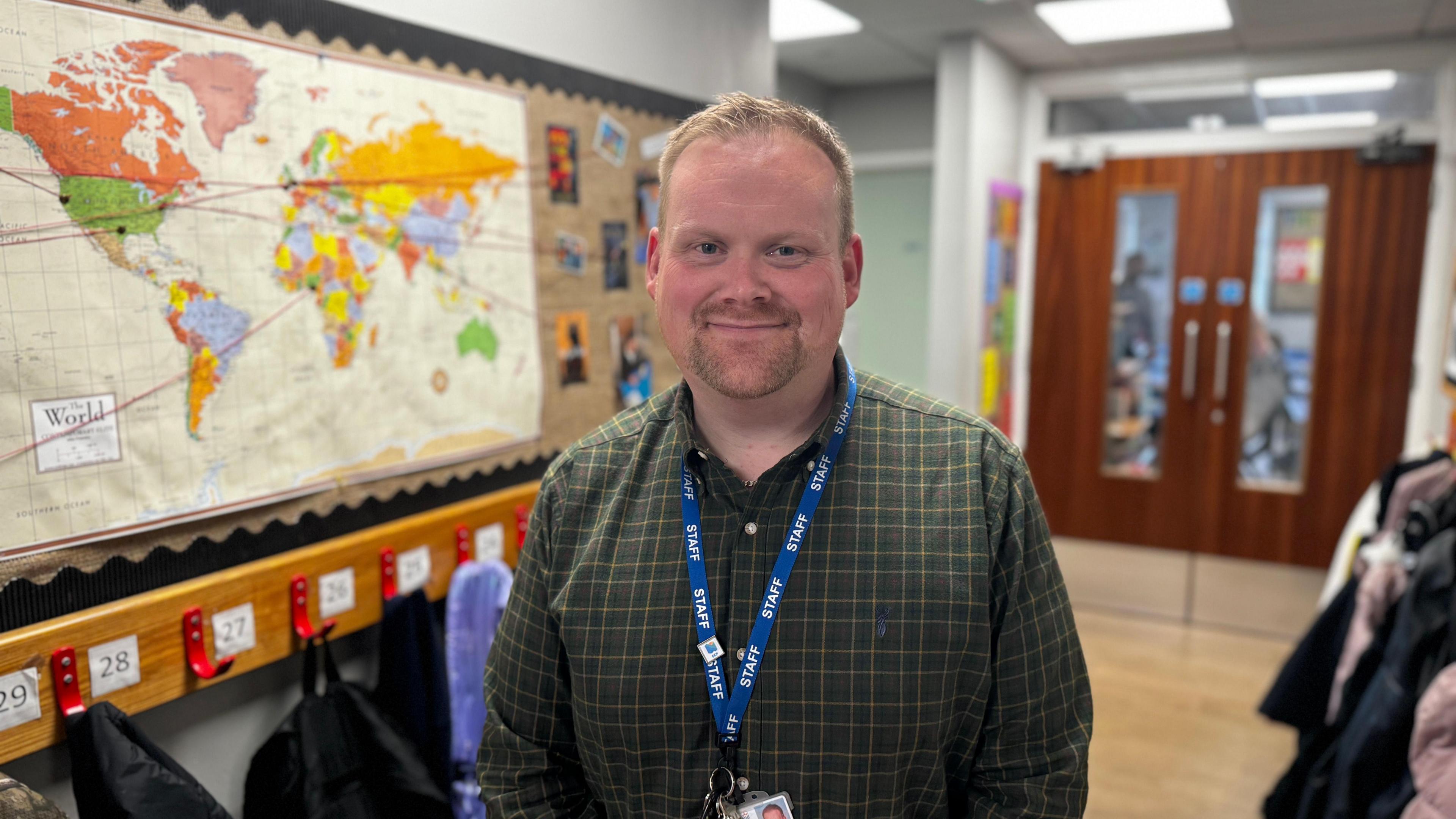 Deputy head teacher Jack Cunningham in the classroom. He wears a dark green shirt and a STAFF lanyard. He has blonde hair. There is world map on his left side.