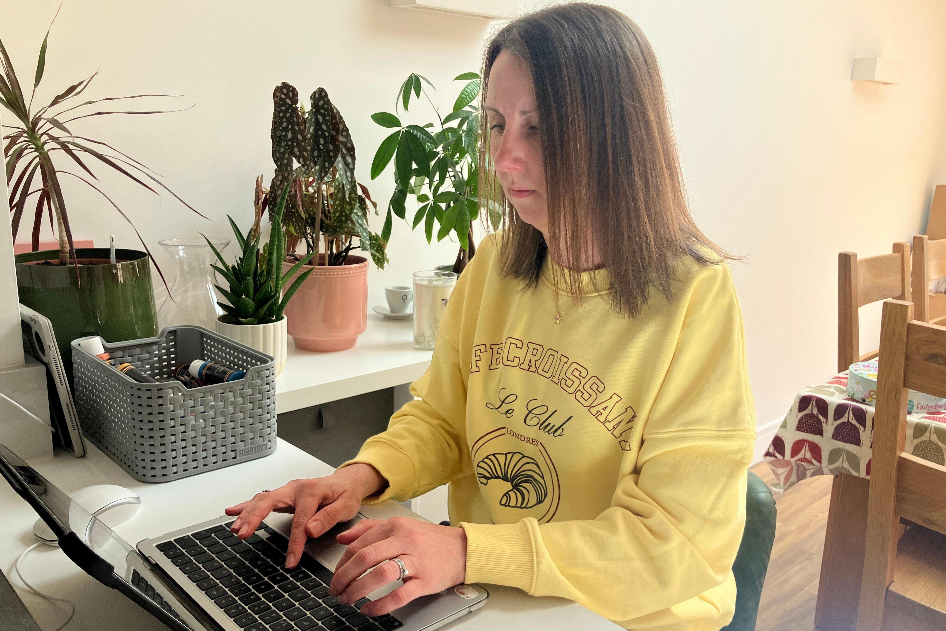 A woman with medium-length brown hair wearing a bright yellow jumper typing on her laptop, she is sat down on a green chair.