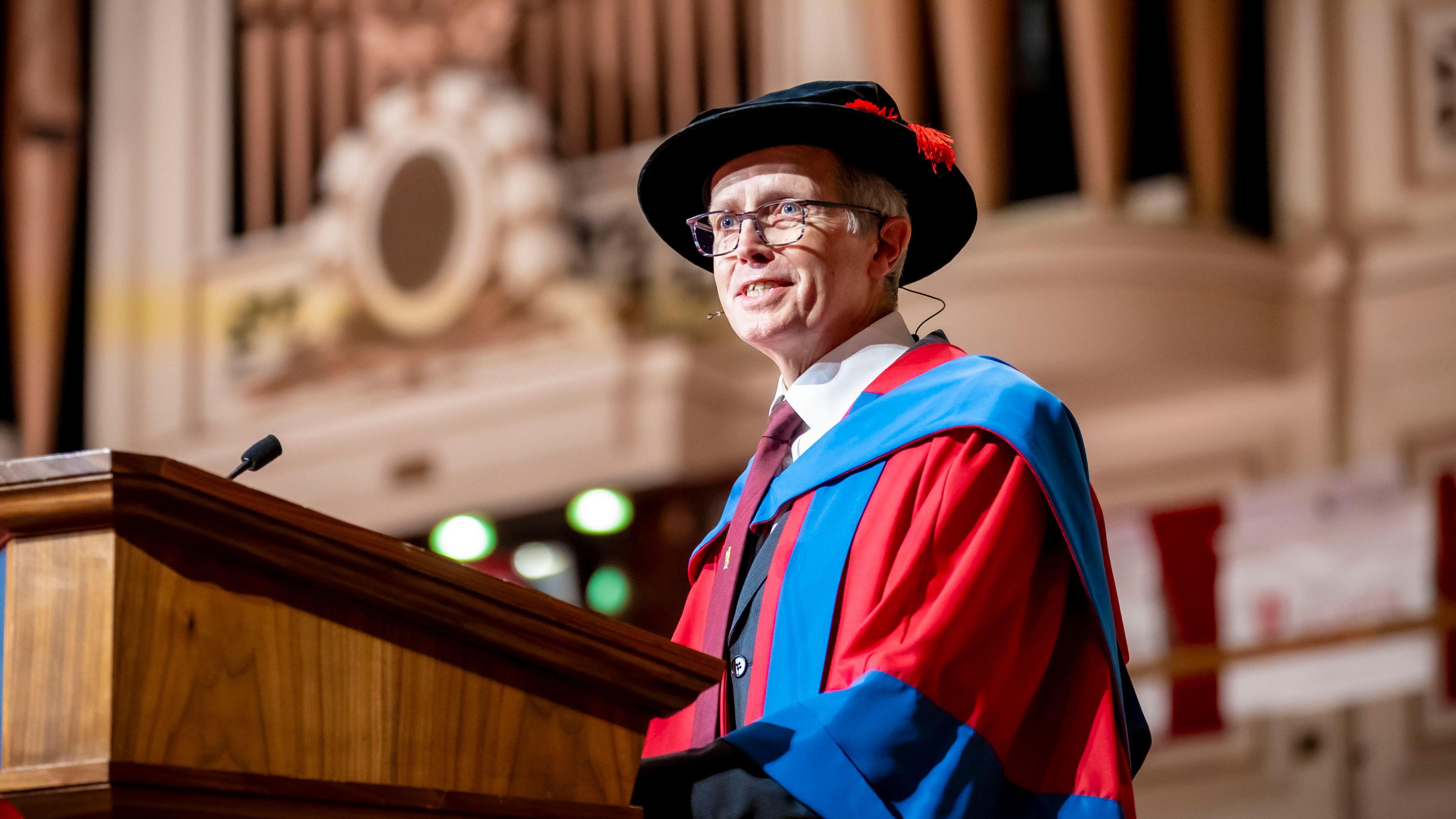 Dr Nigel Hewett wearing a red and blue gown, black hat. He is standing at a wooden podium that has a microphone on top.