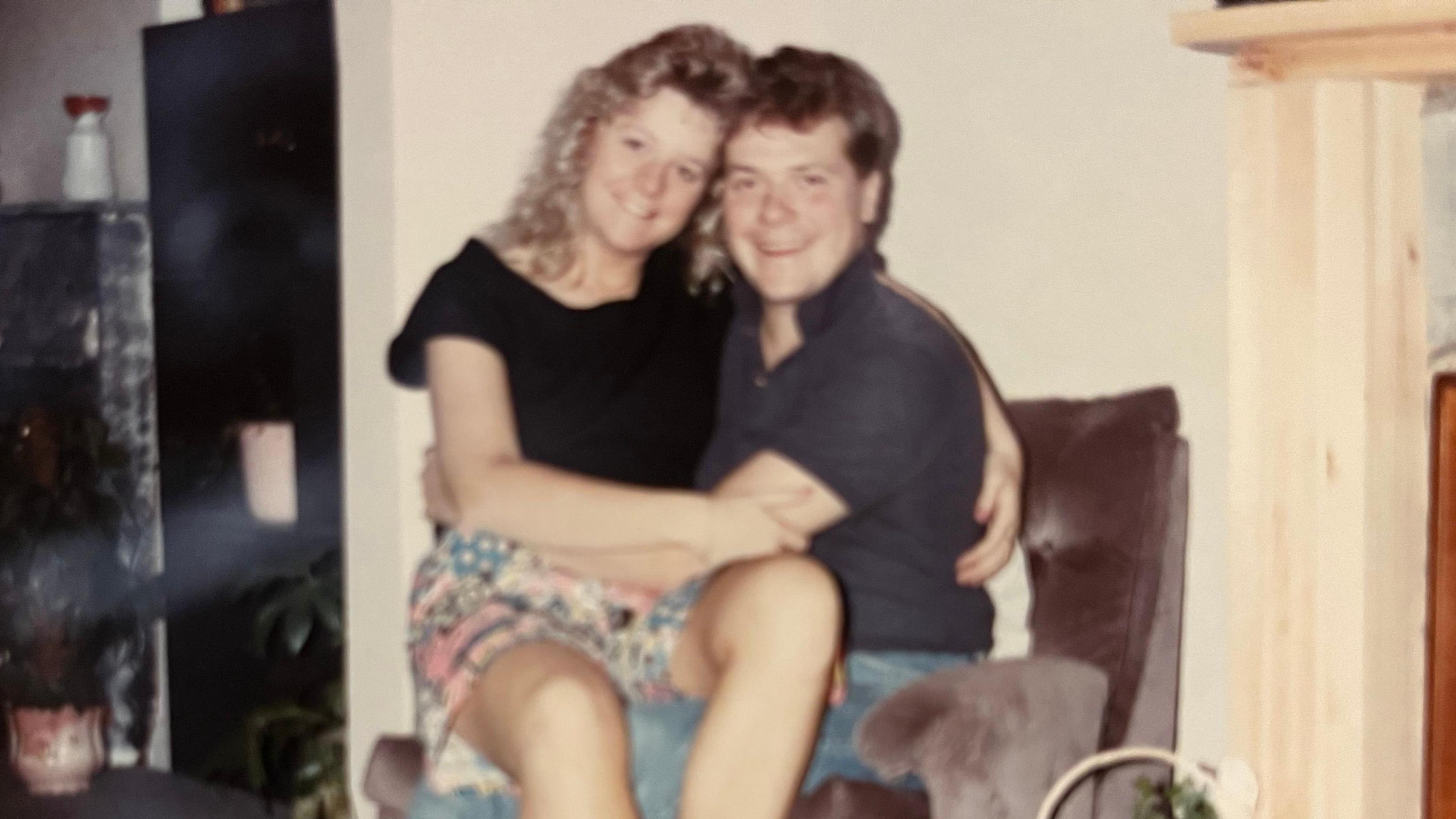 In an old picture, Sue sits on Alex's lap, both smiling. She has curly blonde hair down to her shoulders, wears a black T-shirt and colourful shorts. He sits in a soft brown/grey armchair and they have their arms around each other. He wears a grey/black T-shirt and jeans and they are in what looks like a living room. It is a slightly grainy picture of a picture. 