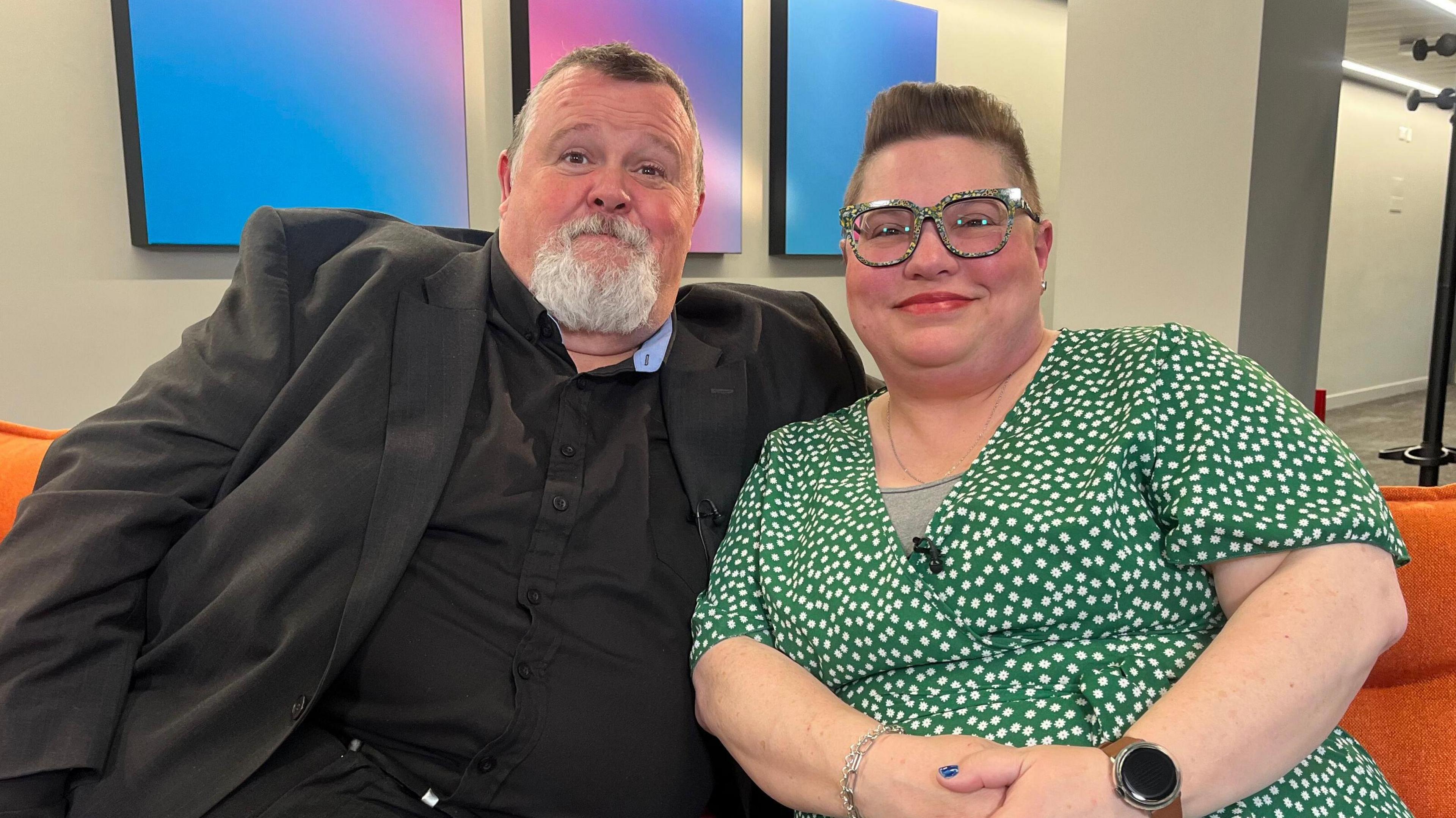 A couple are sitting on an orange sofa. On the left, the man has short dark hair and a gre goatee beard. He is wearing a black shirt and suit jacket. His wife on the right has short hair with an undercut, pattered glasses and a green dress with white spots. They're both smiling.