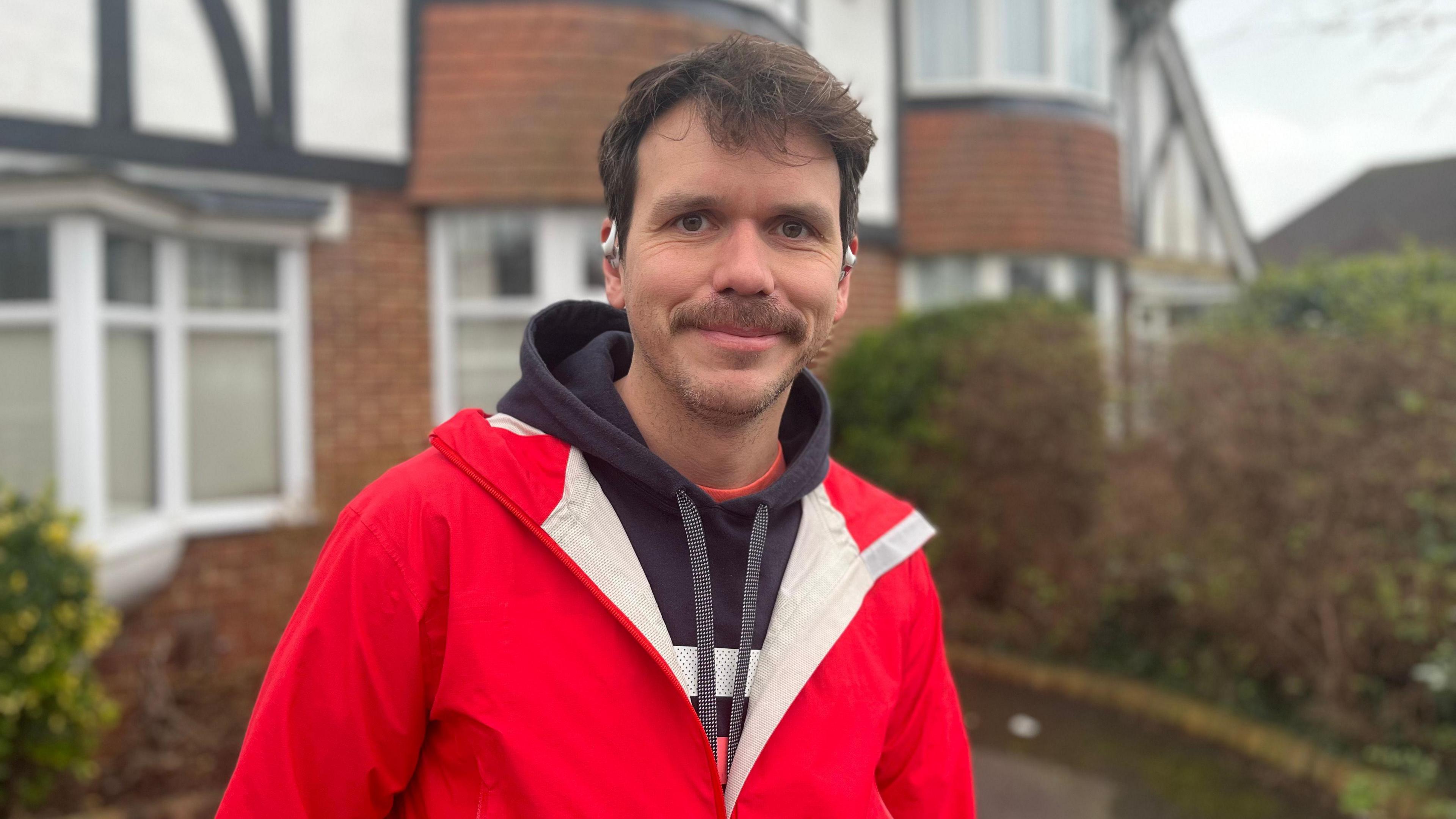 James Roberts, who has brown hair and a moustache. and is wearing a red raincoat and dark hoodie. He is standing outside a house in Hove.