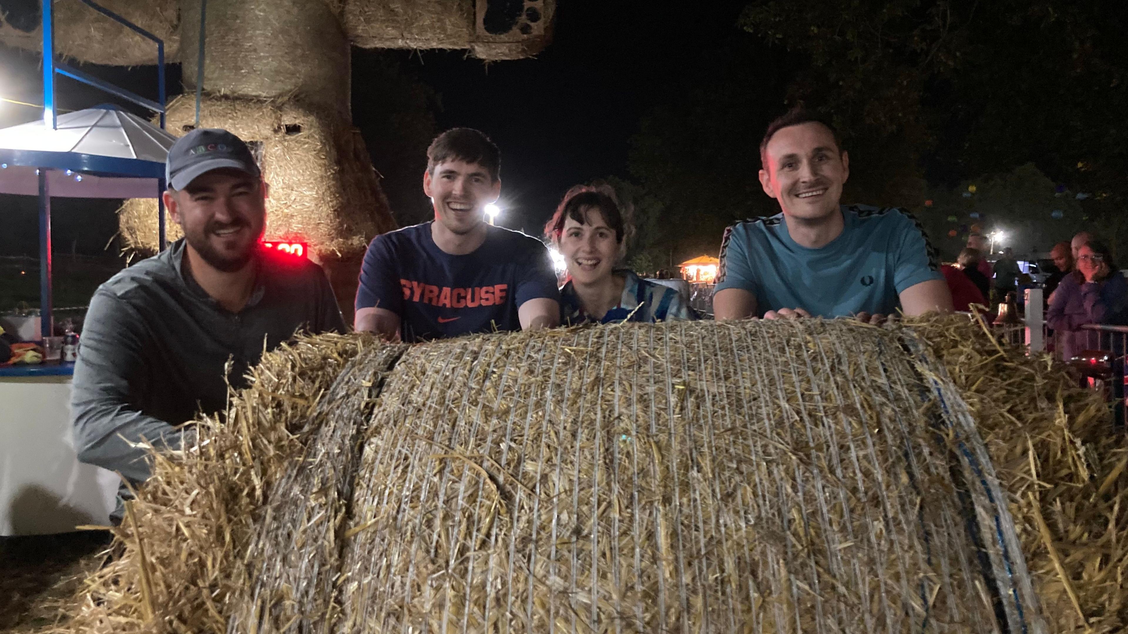 Three men and a woman stand behind a large straw bale, smiling. It is getting dark and a row of lights on string is visible behind them.