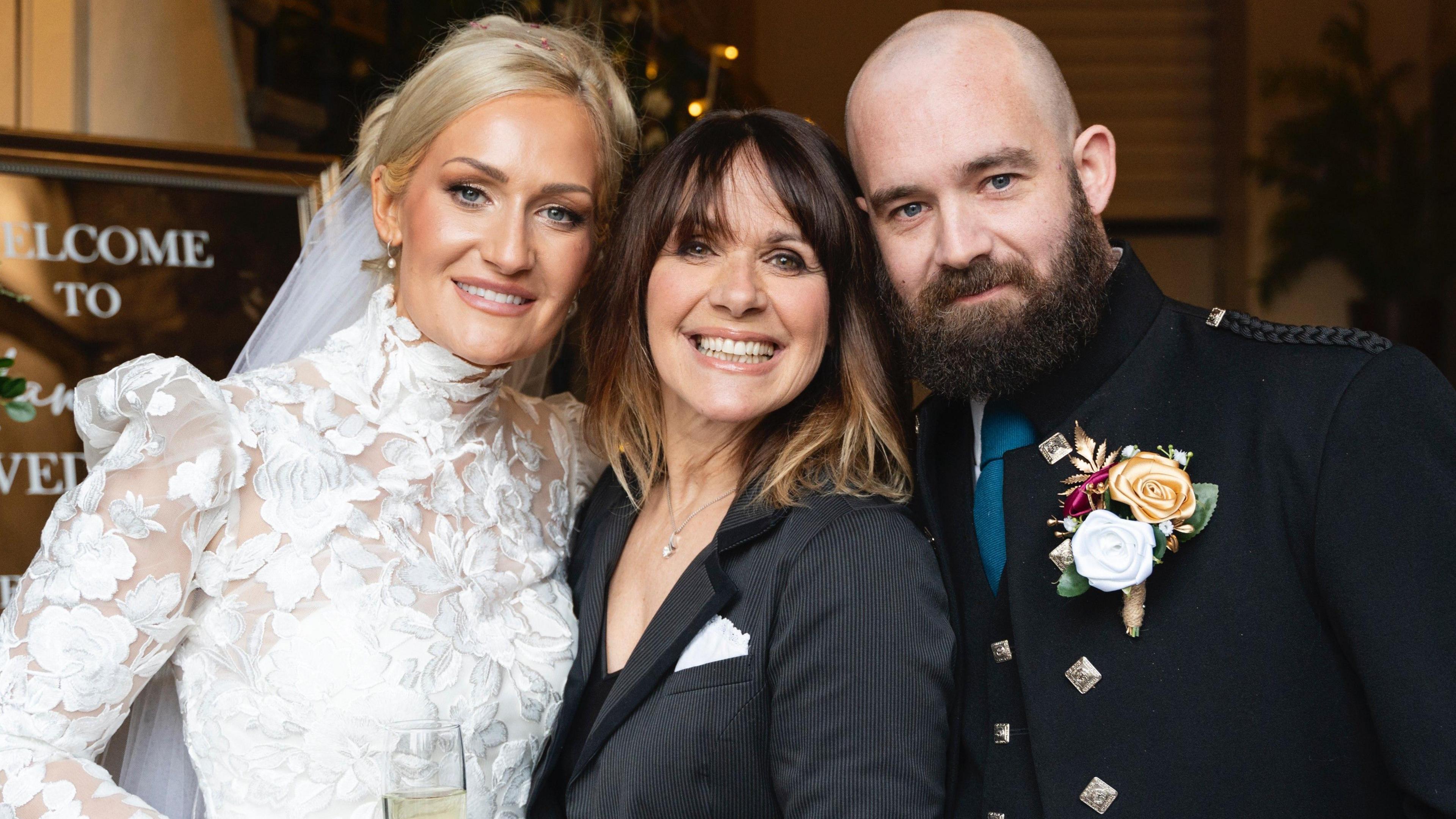 Hannah in a white wedding dress posing alongside Carol Smillie - who has dark hair and is wearing a black jacket - and Hannah's husband, who is bald and wearing a kilt suit jacket. All three are smiling. 