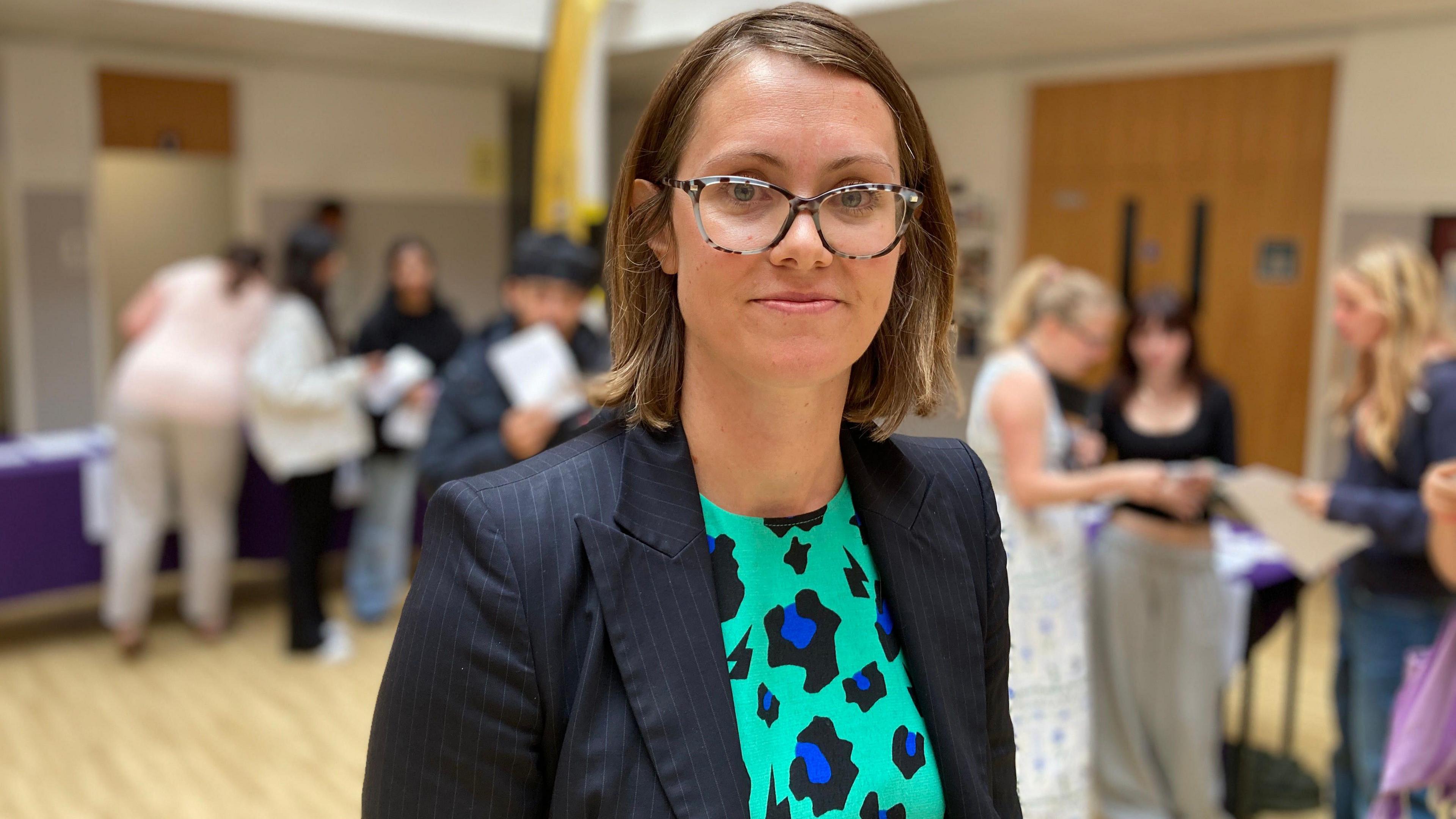Louise Cowley stands in the school hall, with students, parents and teachers gathered in the background. Louise has short light brown hair and wears tortoiseshell thick framed round glasses. She has a blue pinstripe blazer on, with a mint green dress, which has a black and blue leopard print on it. She is smiling at the camera.