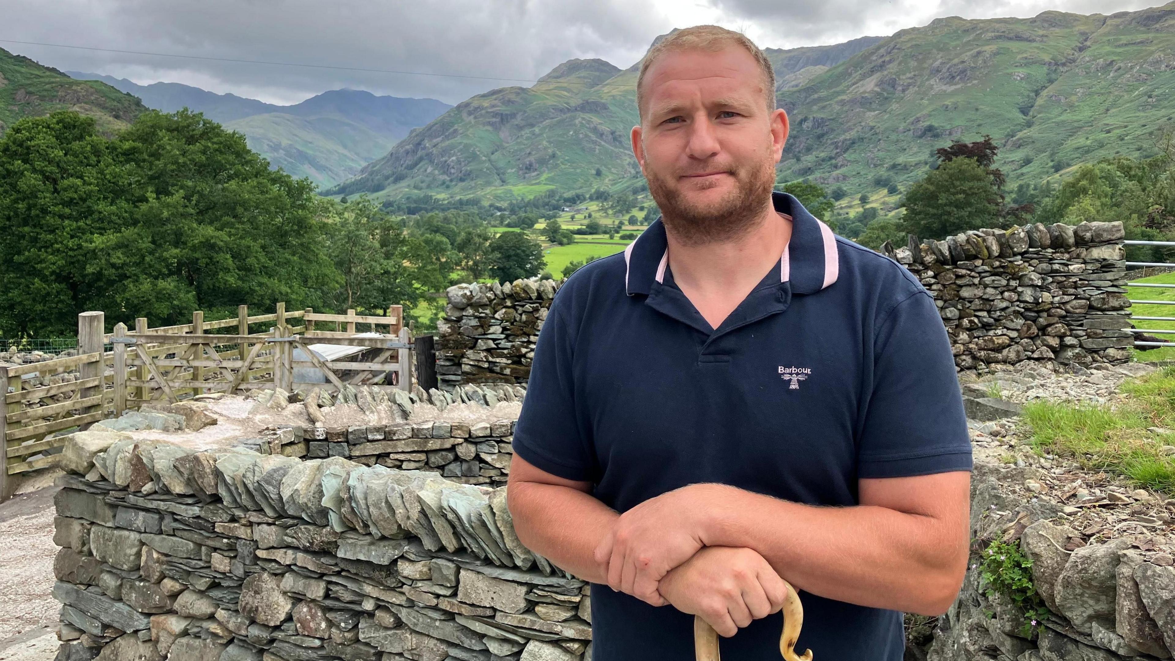 Jonathan Benson in front of a drystone wall at his farm, with the fields and fells in the background. He has short dark blond hair and a beard. He is wearing a blue polo shirt and is holding a wooden walking stick.