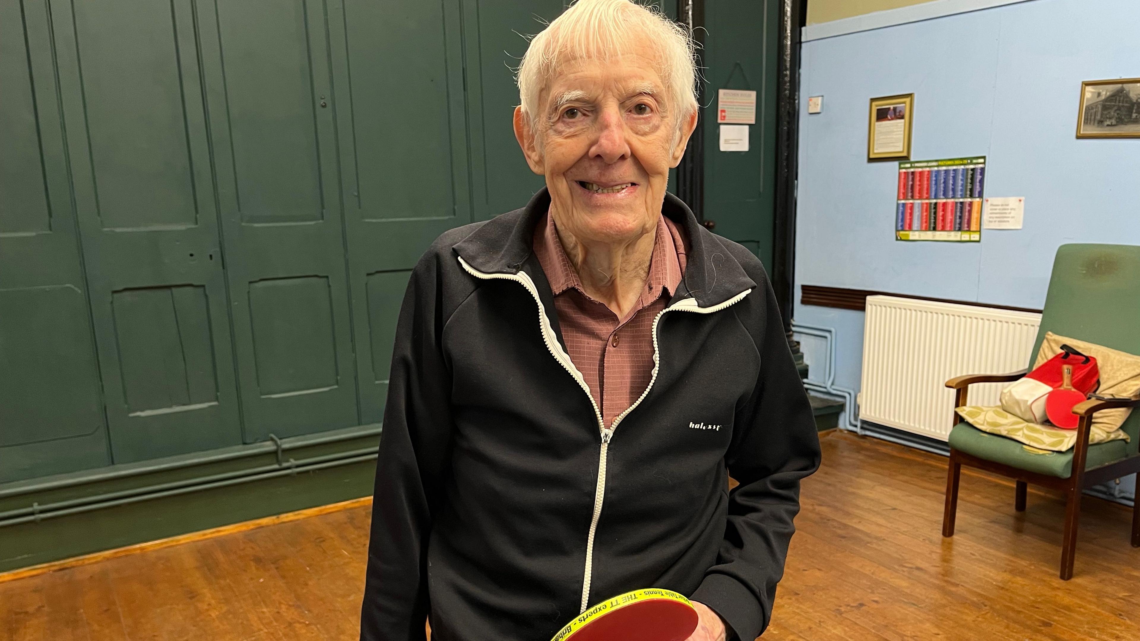 Man in black tracksuit top with grey hair and red shirt holding a red table tennis bat